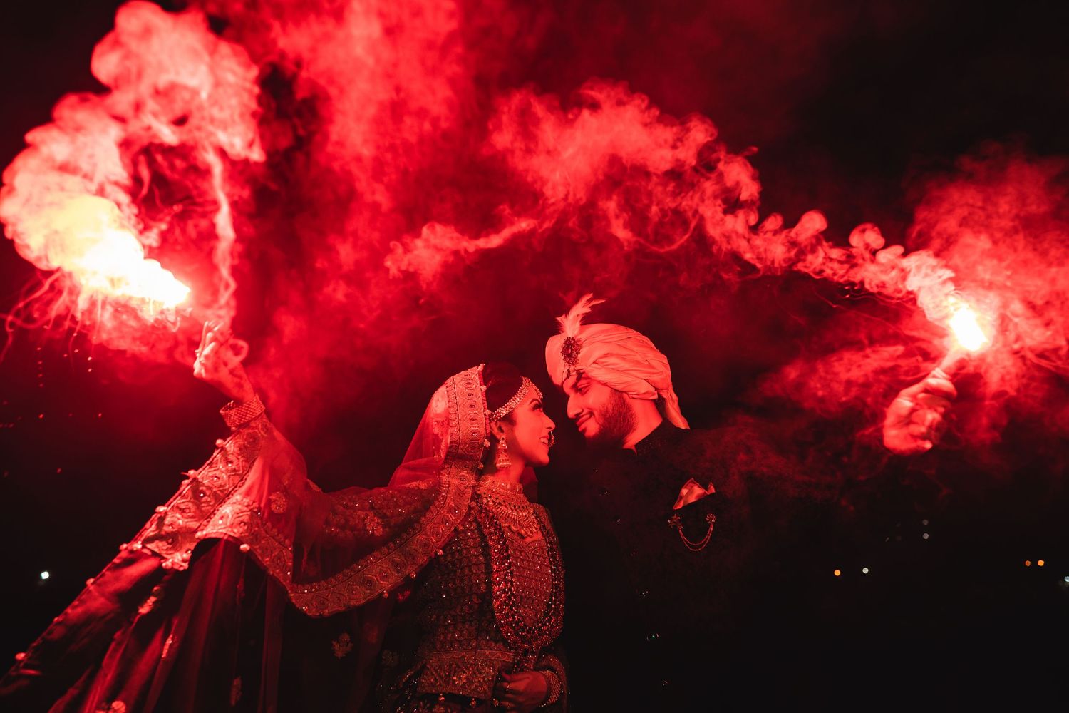 Indian bride and groom holding red smoke flares at night in traditional wedding attire.