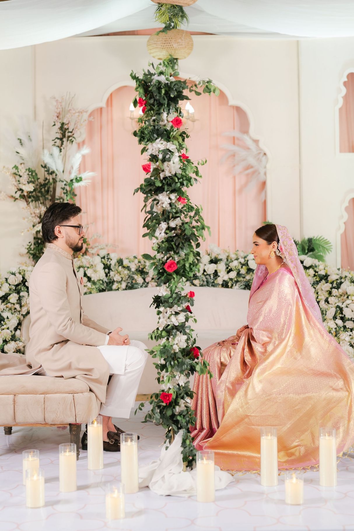 South Asian bride in golden saree and groom in beige sherwani sit together at elegant floral wedding ceremony with candles.
