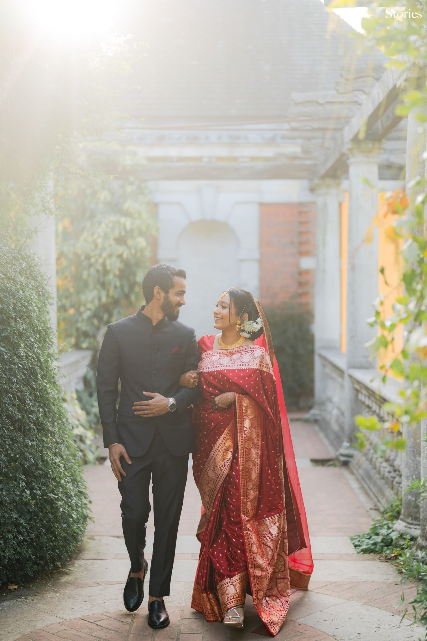 Indian couple in traditional wedding attire walking through elegant garden, bride in red saree, groom in black suit.
