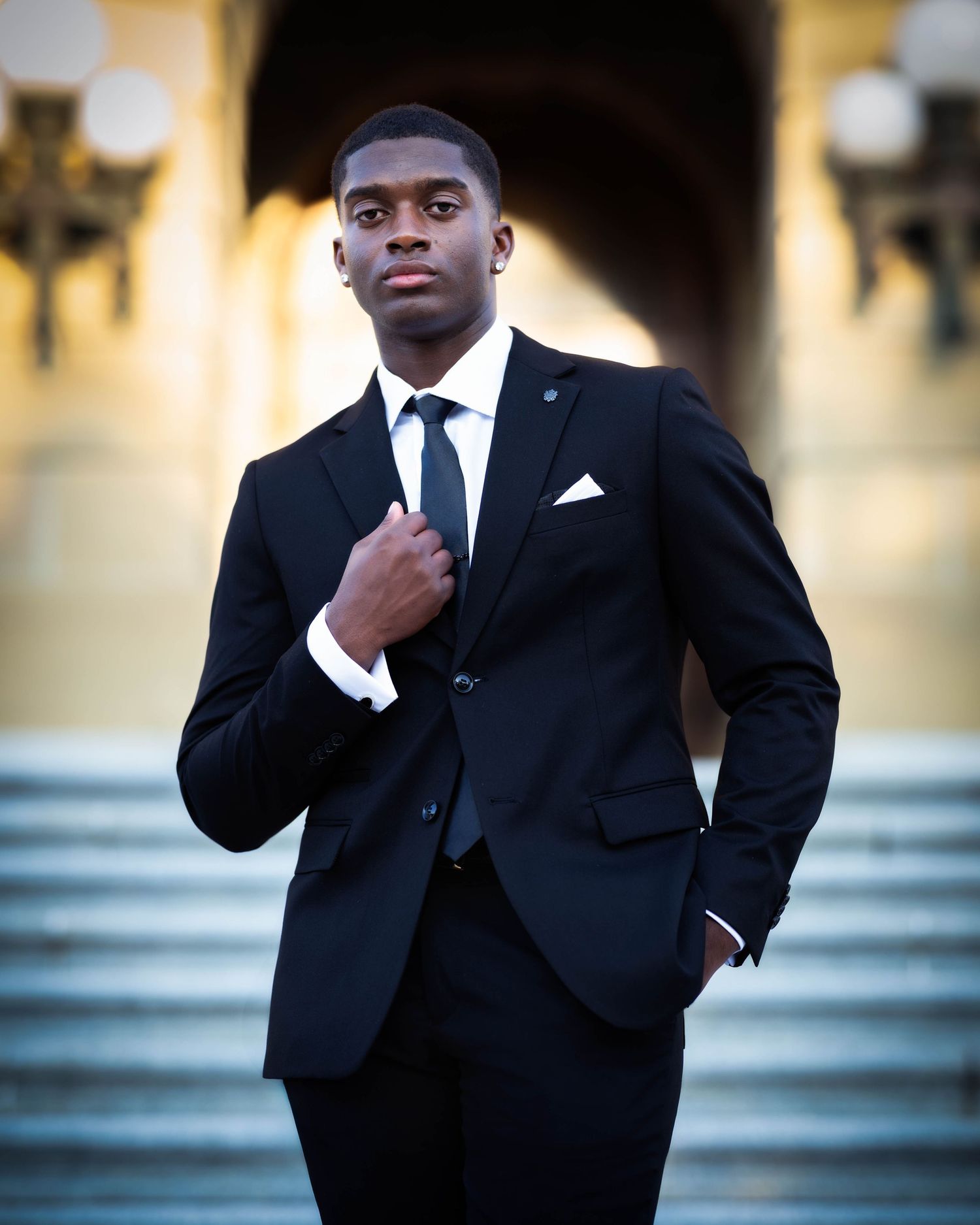 A man in a black suit and tie poses confidently against a blurred background in a professional photo series.