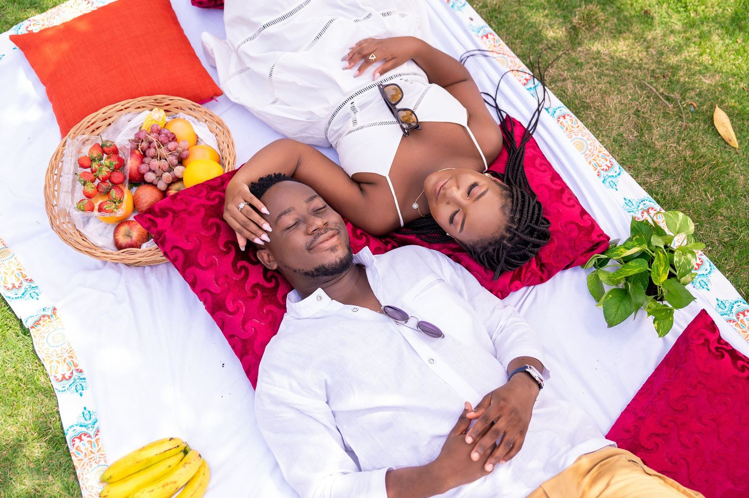 A couple enjoying a romantic picnic on a red blanket with fresh fruit and snacks spread out on the grass.