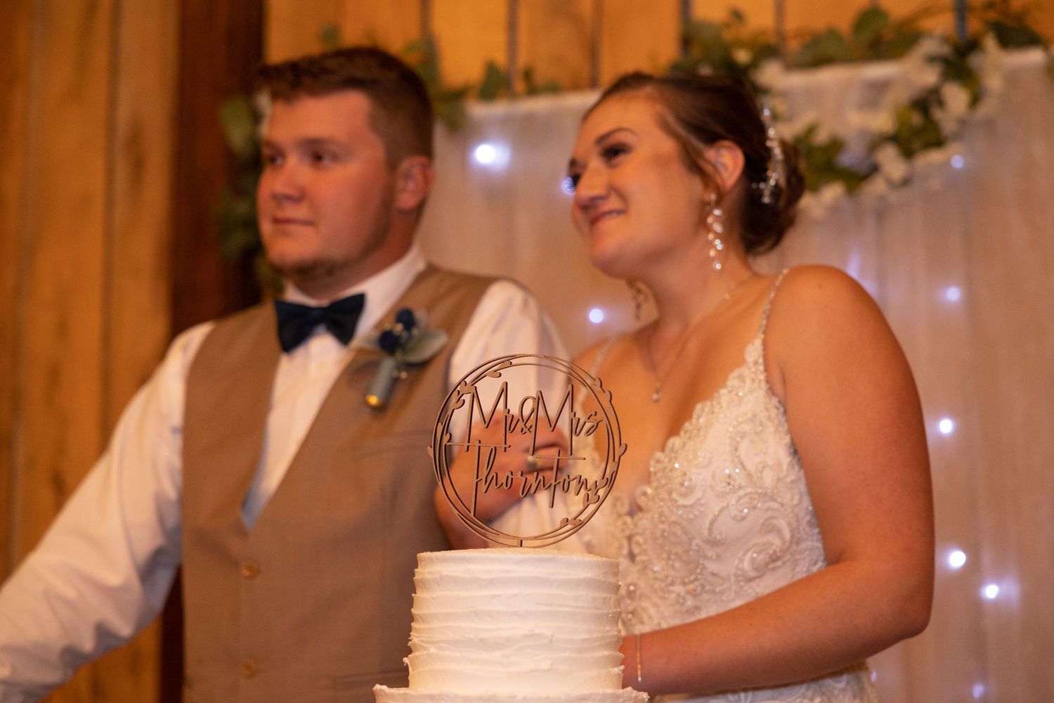 Bride and groom smile beside white wedding cake with decorative topper at rustic barn wedding reception.