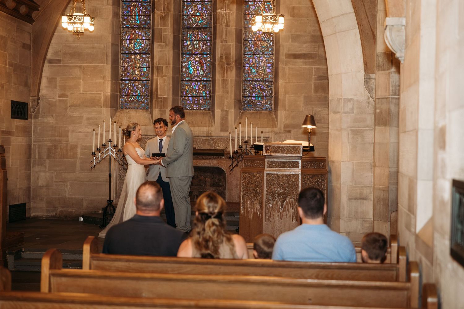 Couple exchanging vows at a stone church altar with stained glass windows and wooden pews filled with guests.