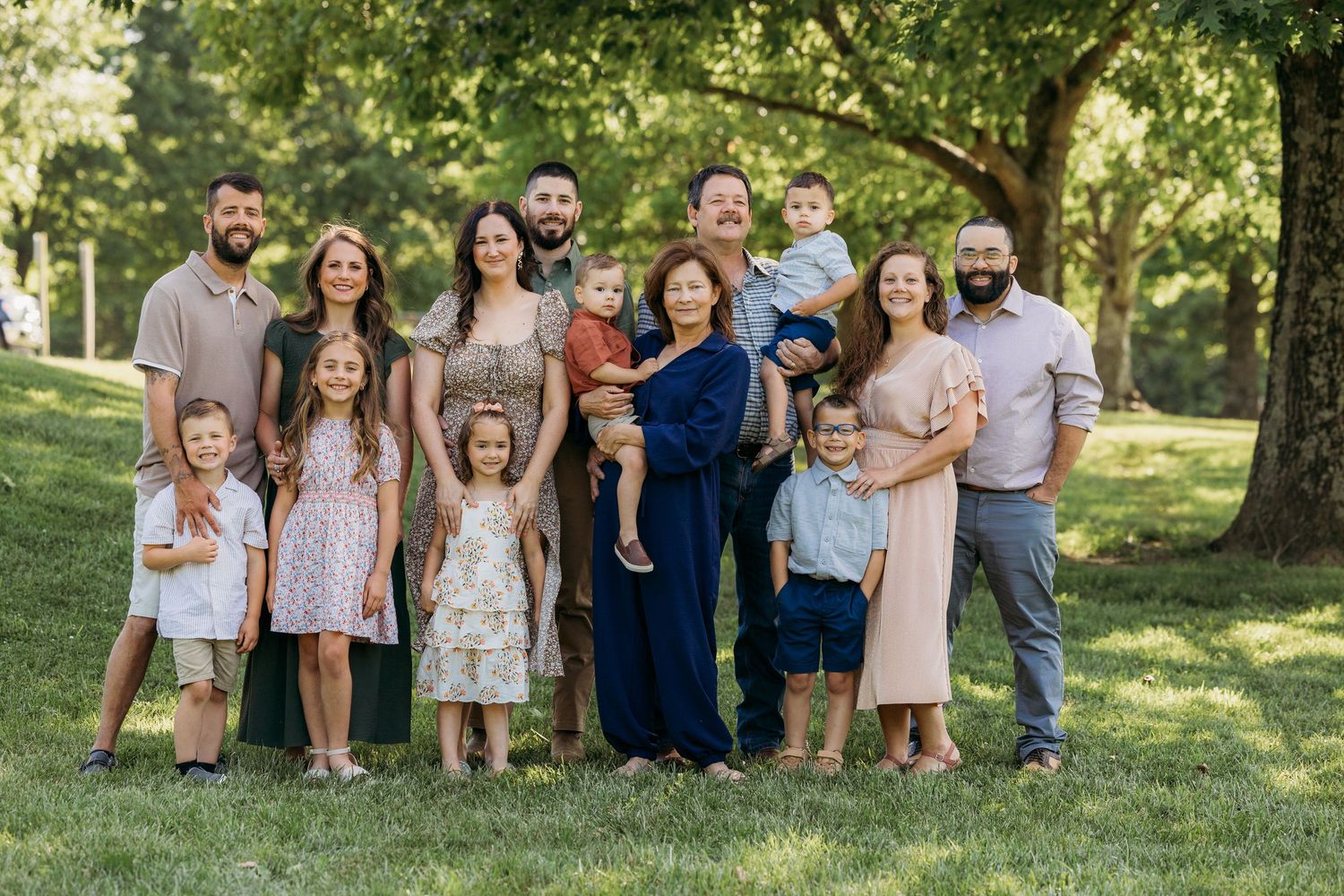 Large multigenerational family portrait outdoors on green grass under shady trees, featuring adults and young children smiling.