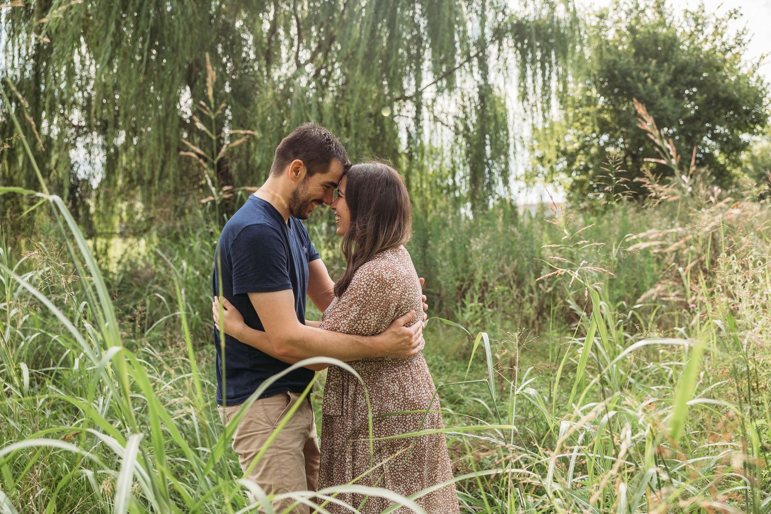 Couple embracing intimately among tall green grasses and weeping willow trees in a lush outdoor setting.