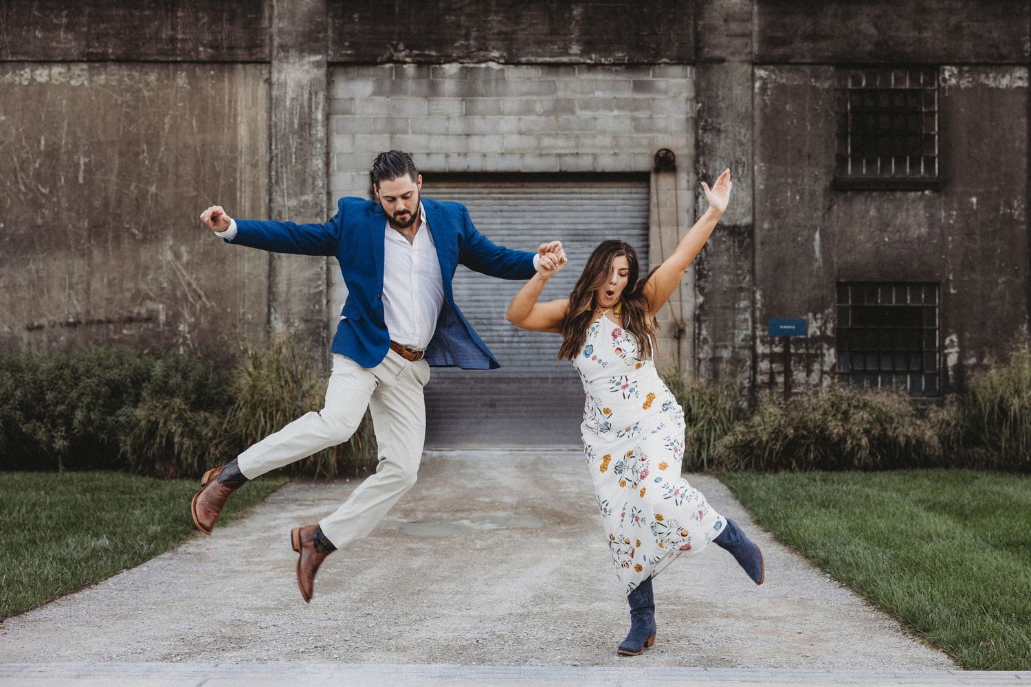 Joyful couple jumping and celebrating outdoors near rustic concrete wall, man in blue blazer, woman in floral dress.