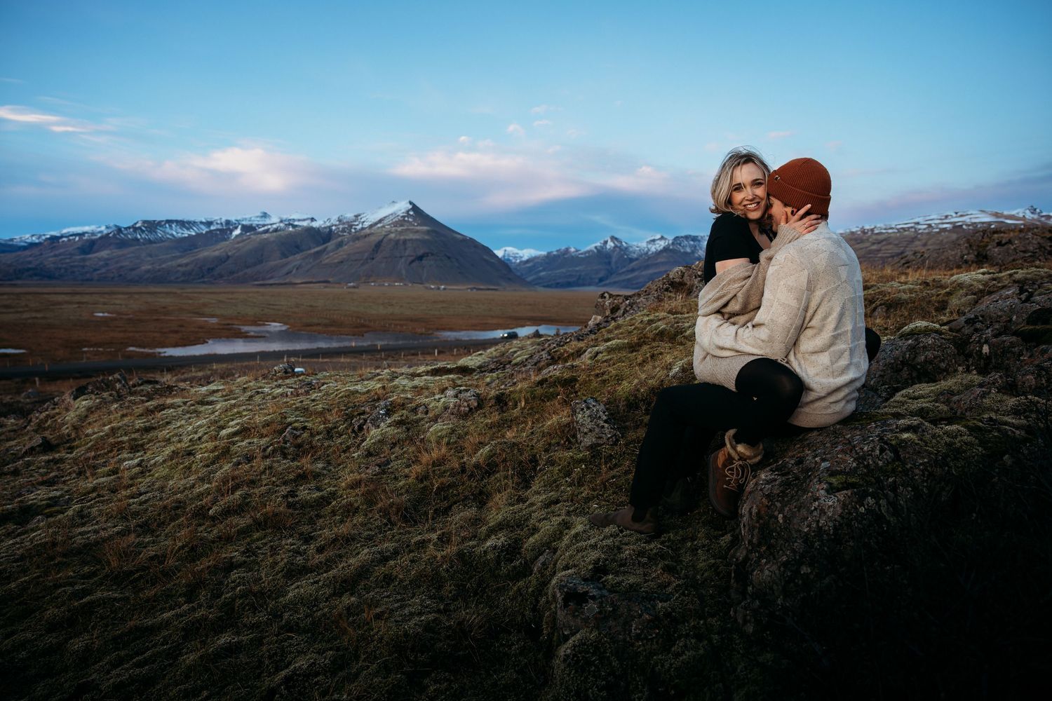 Couple sitting on mossy rocks in Iceland with dramatic mountain landscape and twilight sky backdrop.