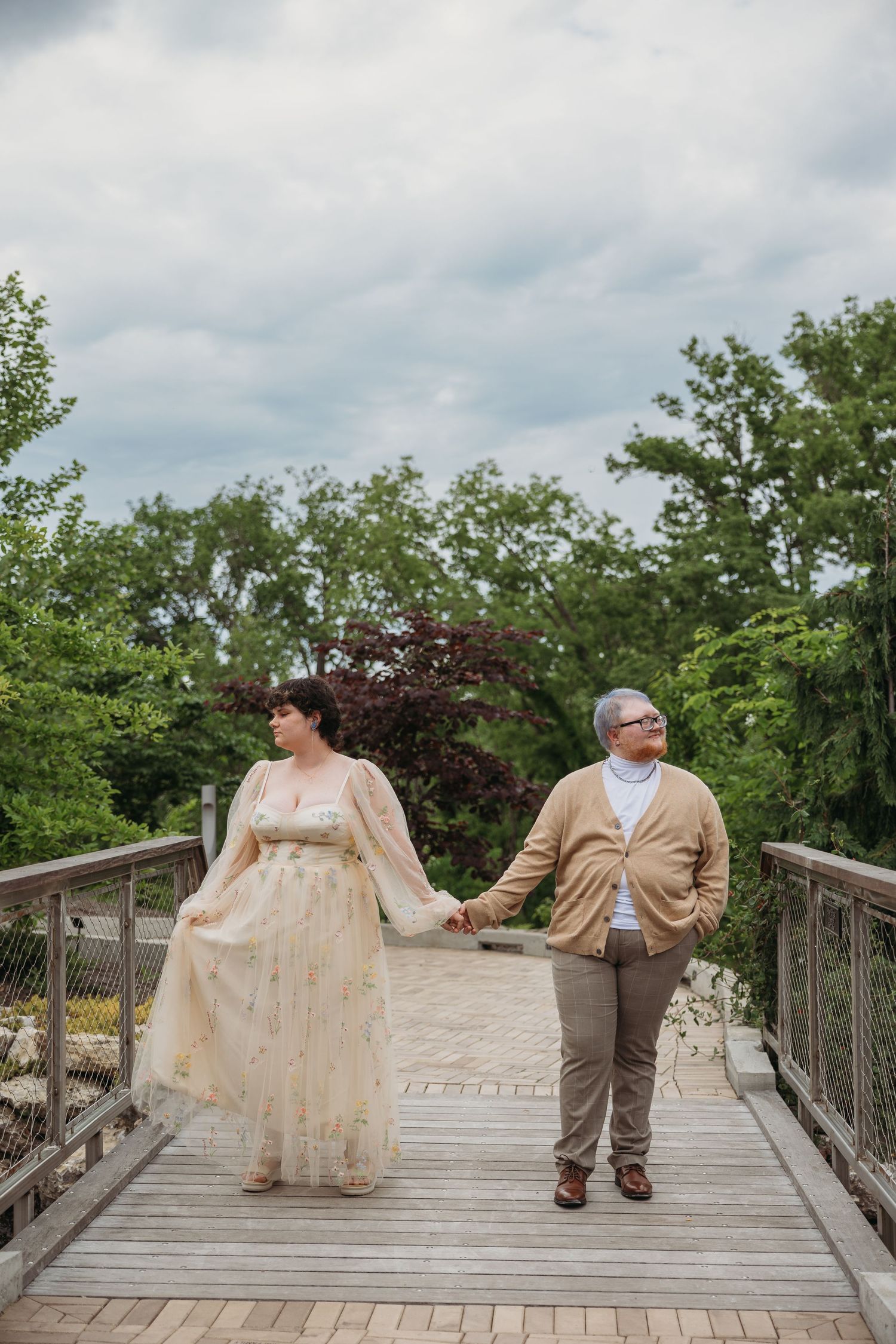 Two brides hold hands on a wooden bridge surrounded by lush green trees on a cloudy day.