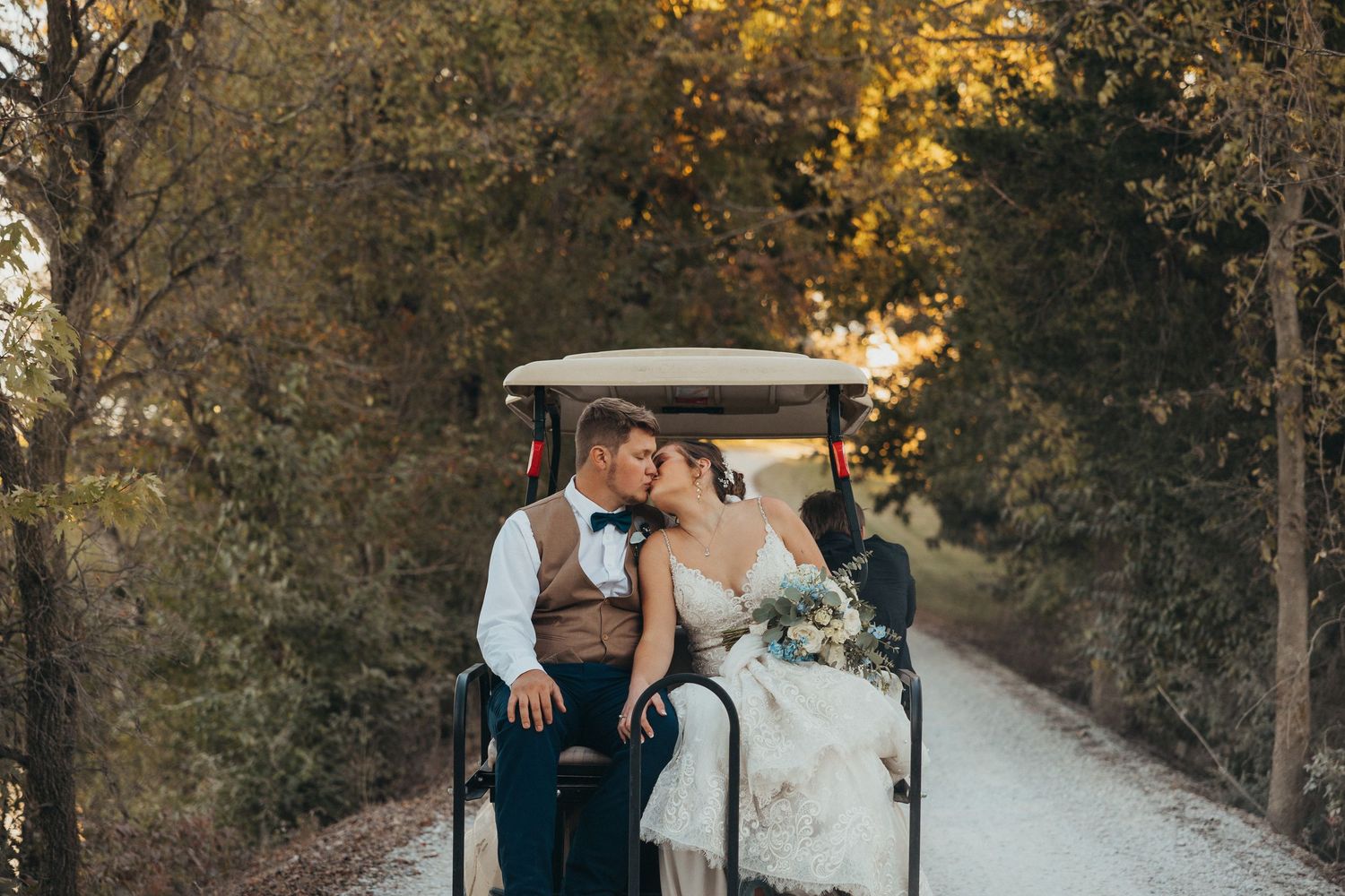 Bride and groom sharing a kiss while riding a golf cart down a tree-lined path at sunset.
