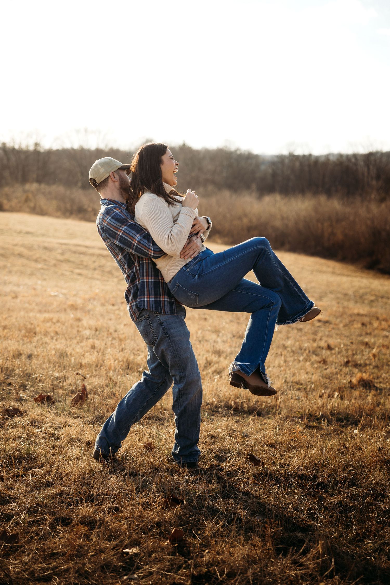 Man lifting woman in his arms in a sunny autumn field, both wearing jeans and flannel, laughing joyfully.