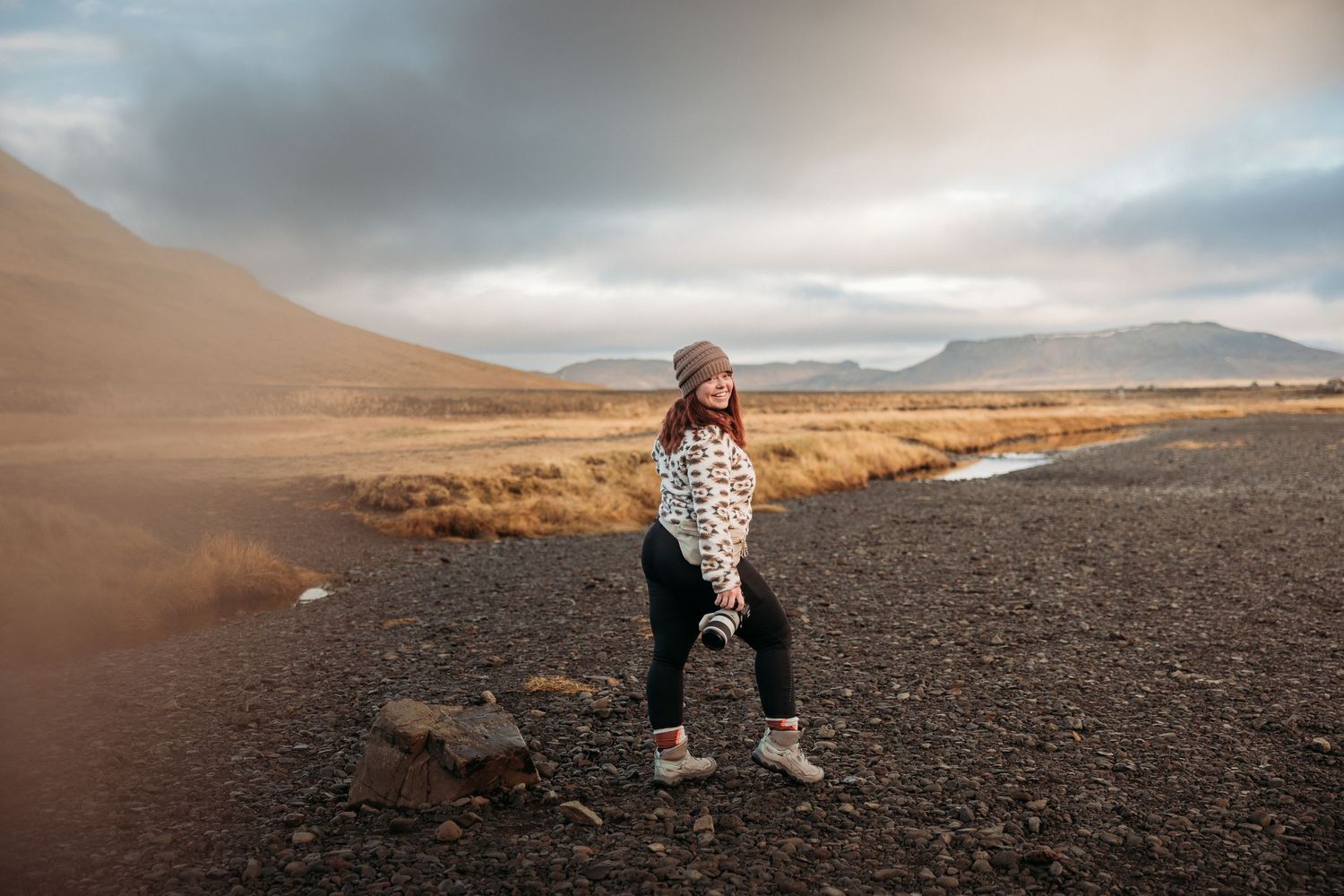 Woman in patterned sweater and beanie poses on volcanic landscape with mountains and dramatic cloudy sky in Iceland.