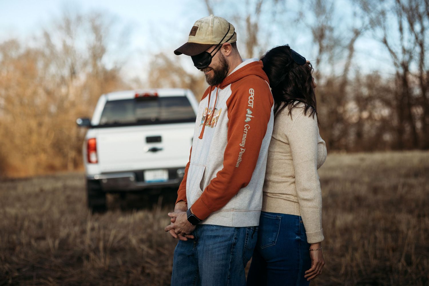 Couple holding hands standing in a field near a white pickup truck during autumn.