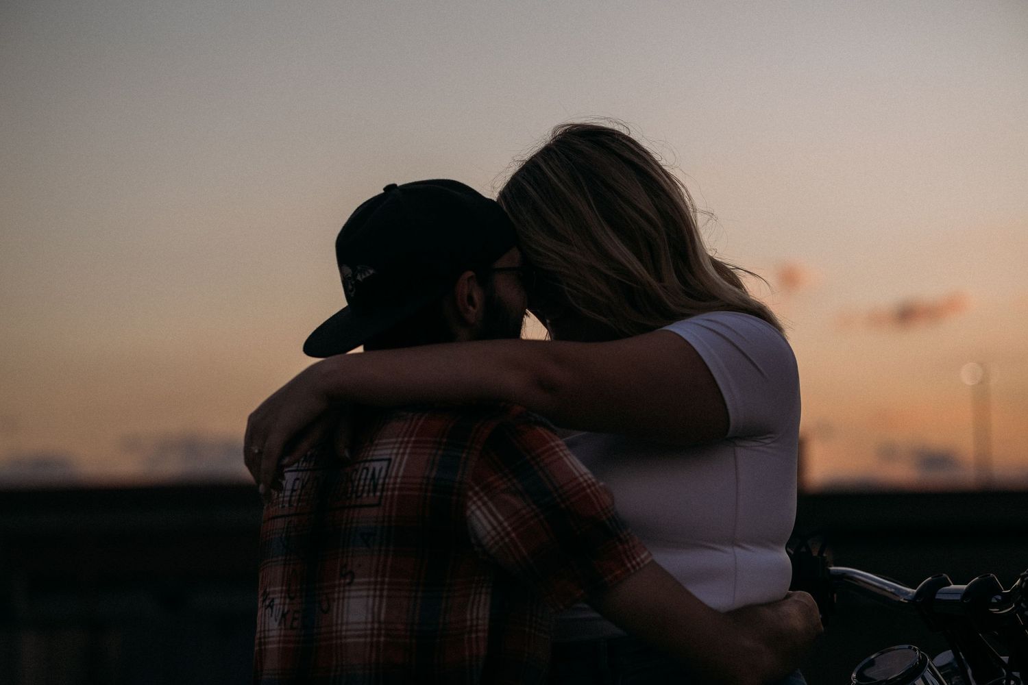 Couple embracing at sunset near a motorcycle, sharing a romantic moment in warm golden hour light.