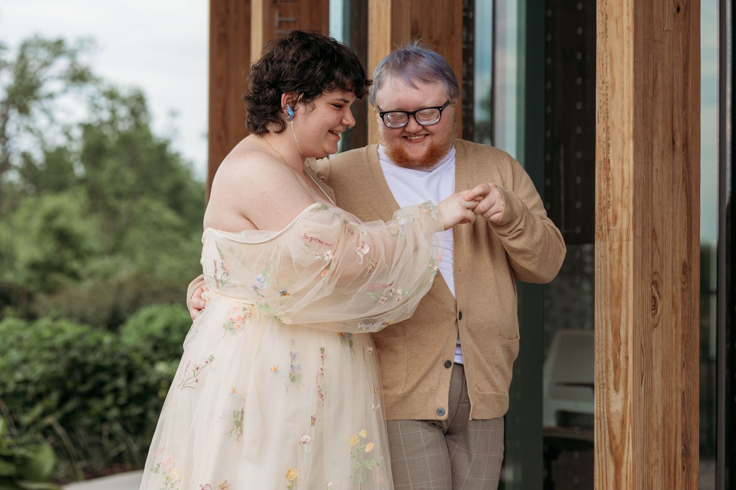 A joyful couple in wedding attire laughing together outdoors, bride in floral tulle gown, groom in tan cardigan.
