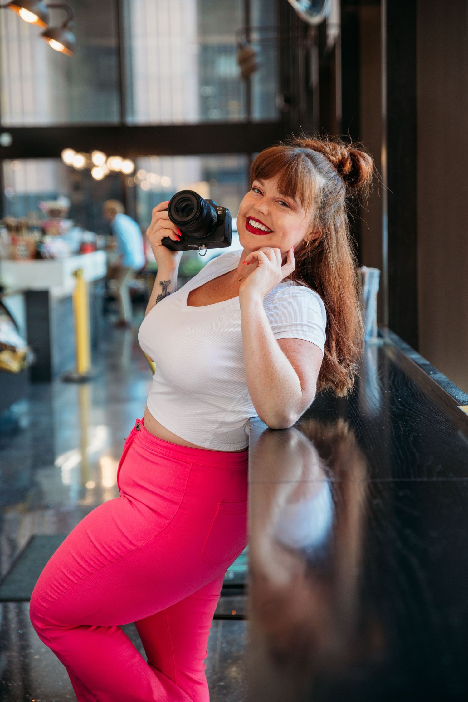 Smiling woman in white crop top and pink pants holding a DSLR camera, leaning on a counter in a modern cafe.