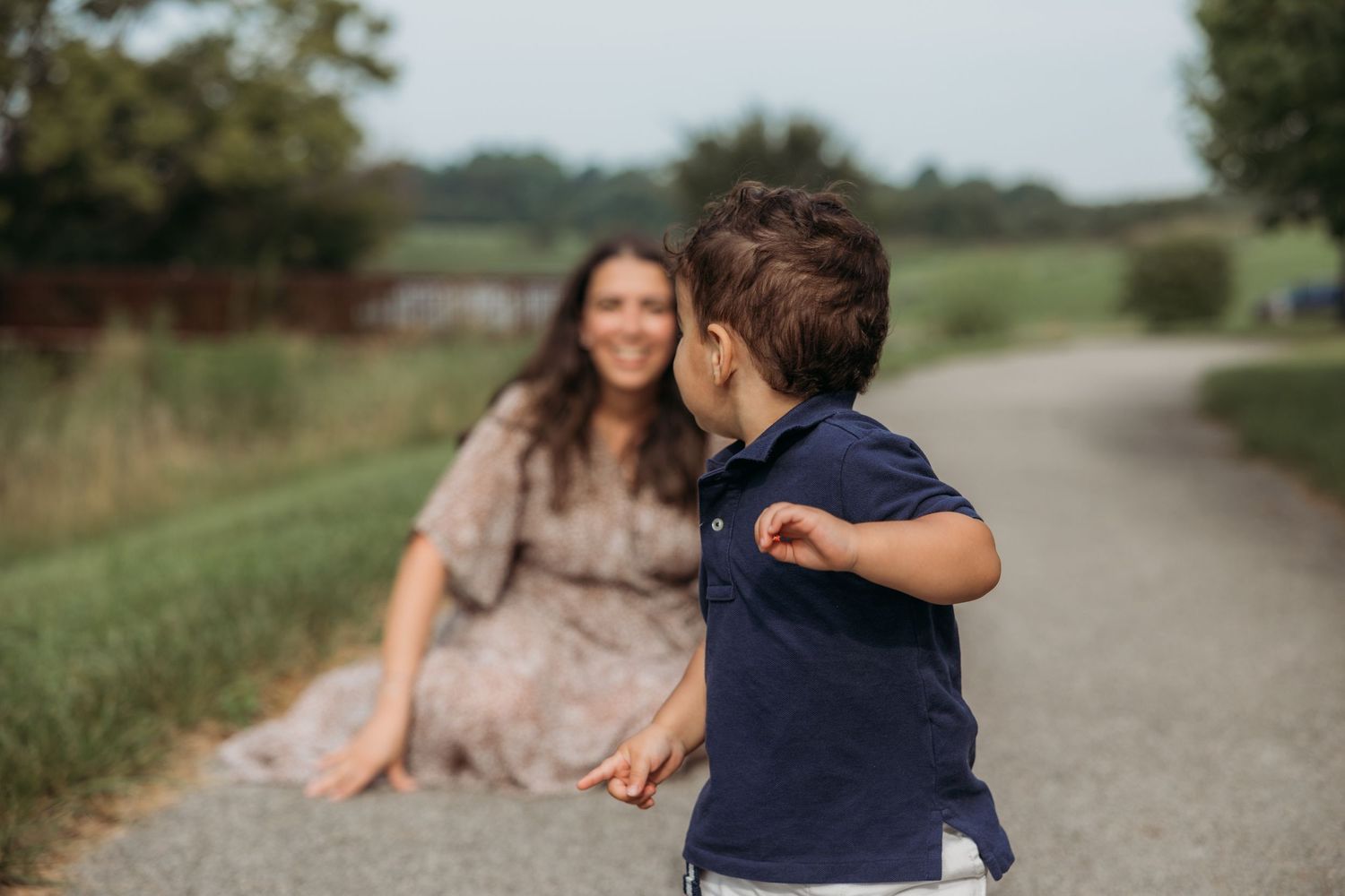 A young boy runs toward his smiling mother sitting on a path outdoors during a family photo session.