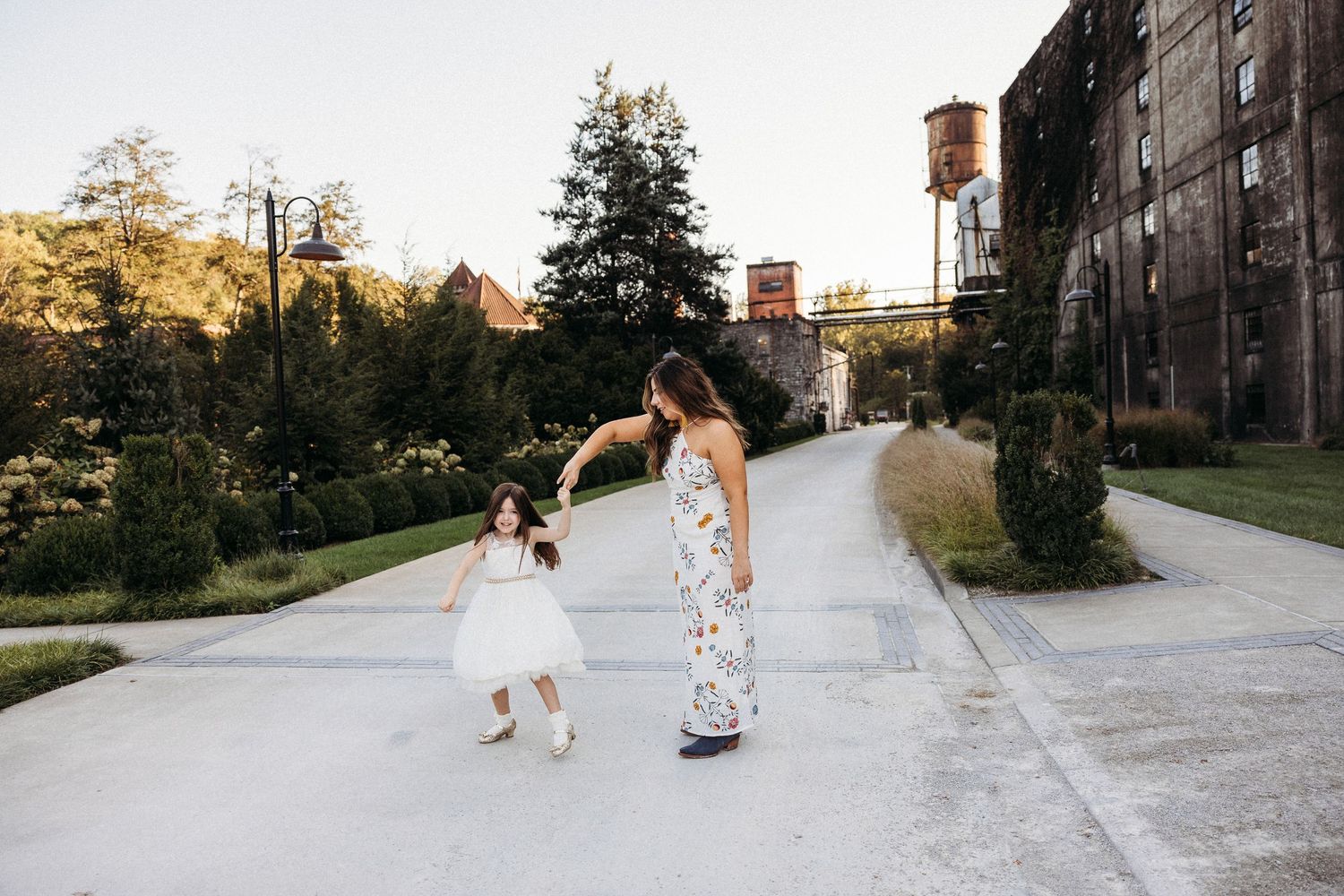 A mother and young daughter in white dresses dance together on a pathway near a historic brick building at sunset.
