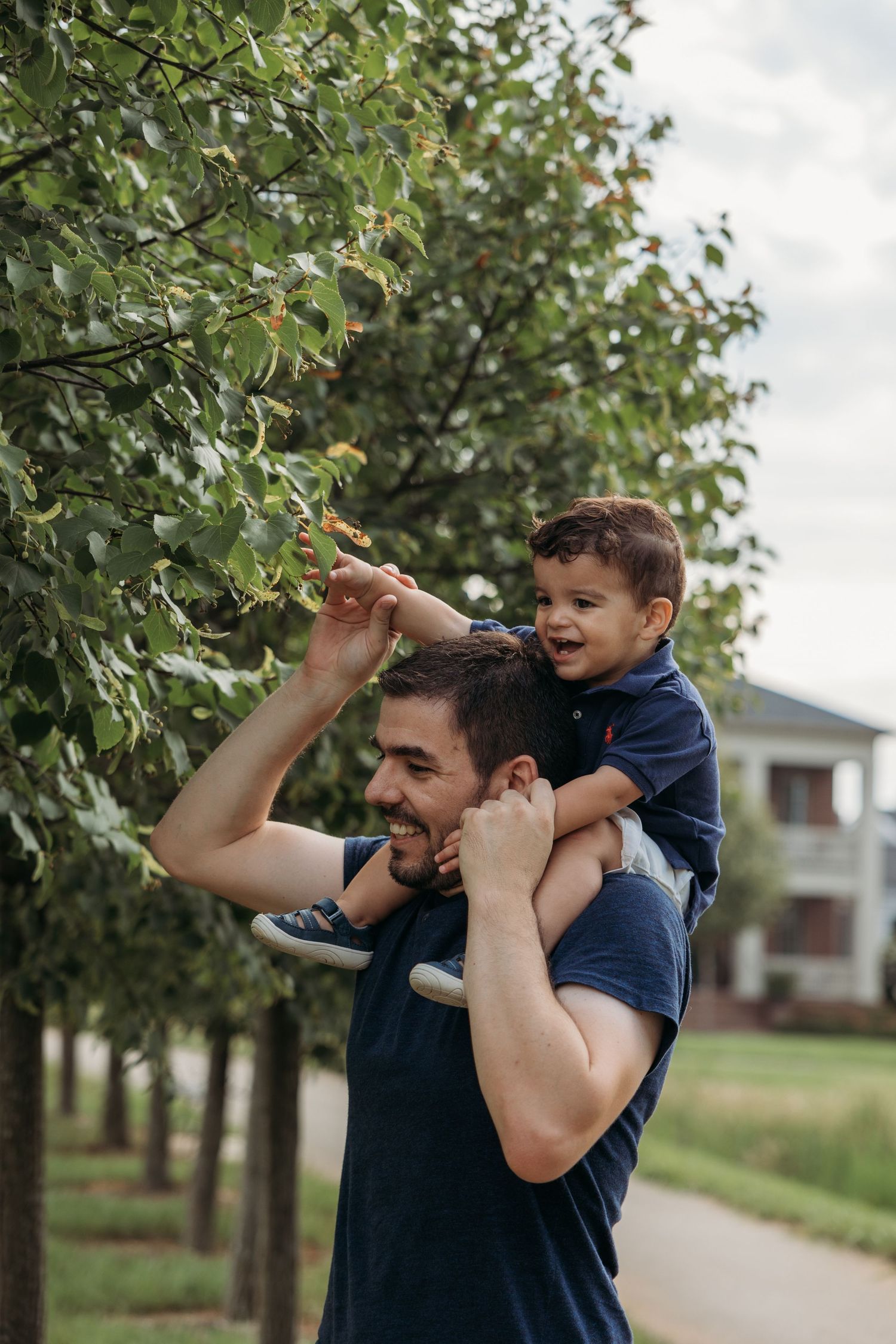 A smiling father carries his laughing toddler son on his shoulders while reaching for tree branches in a park.