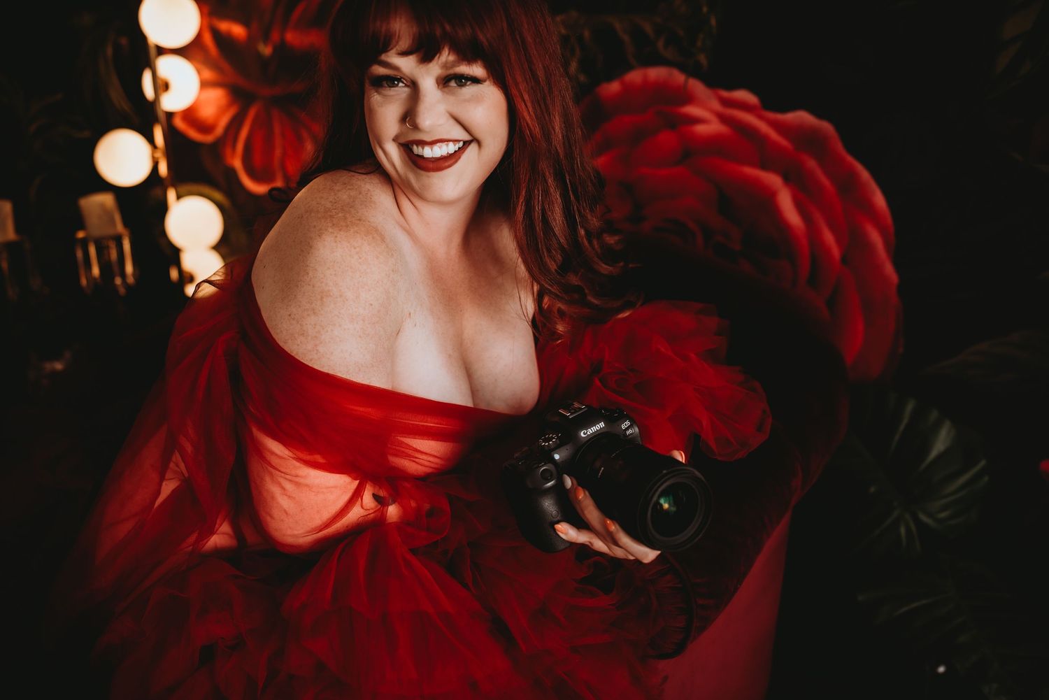 Woman in red tulle gown holding a camera, smiling warmly, surrounded by warm bokeh lights.