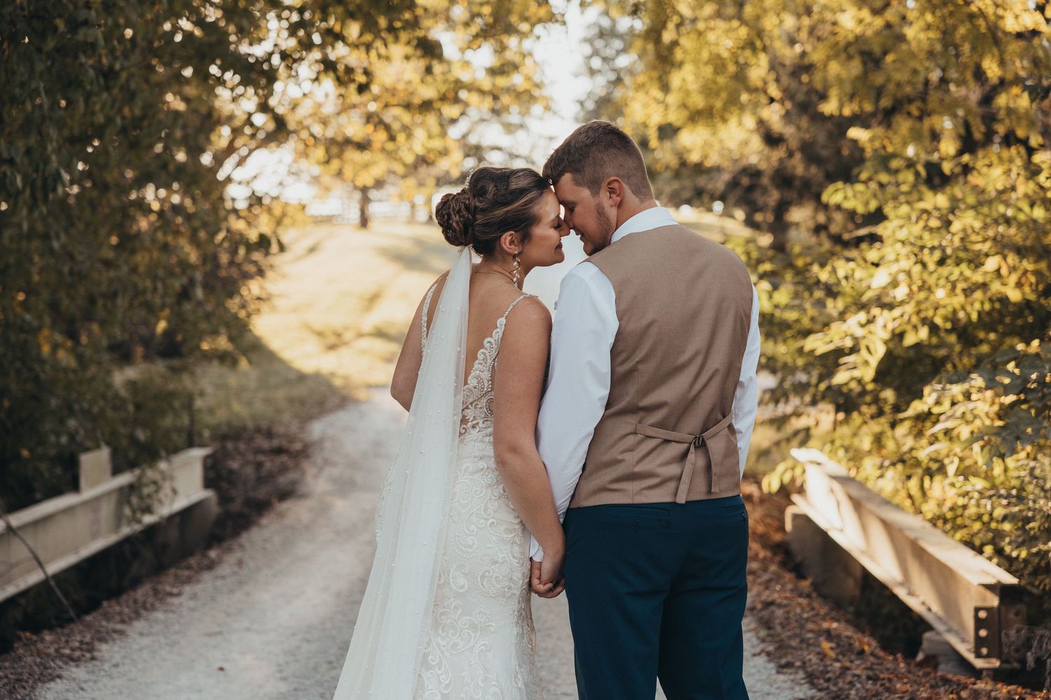Bride and groom touching foreheads while holding hands on tree-lined path in romantic wedding portrait.
