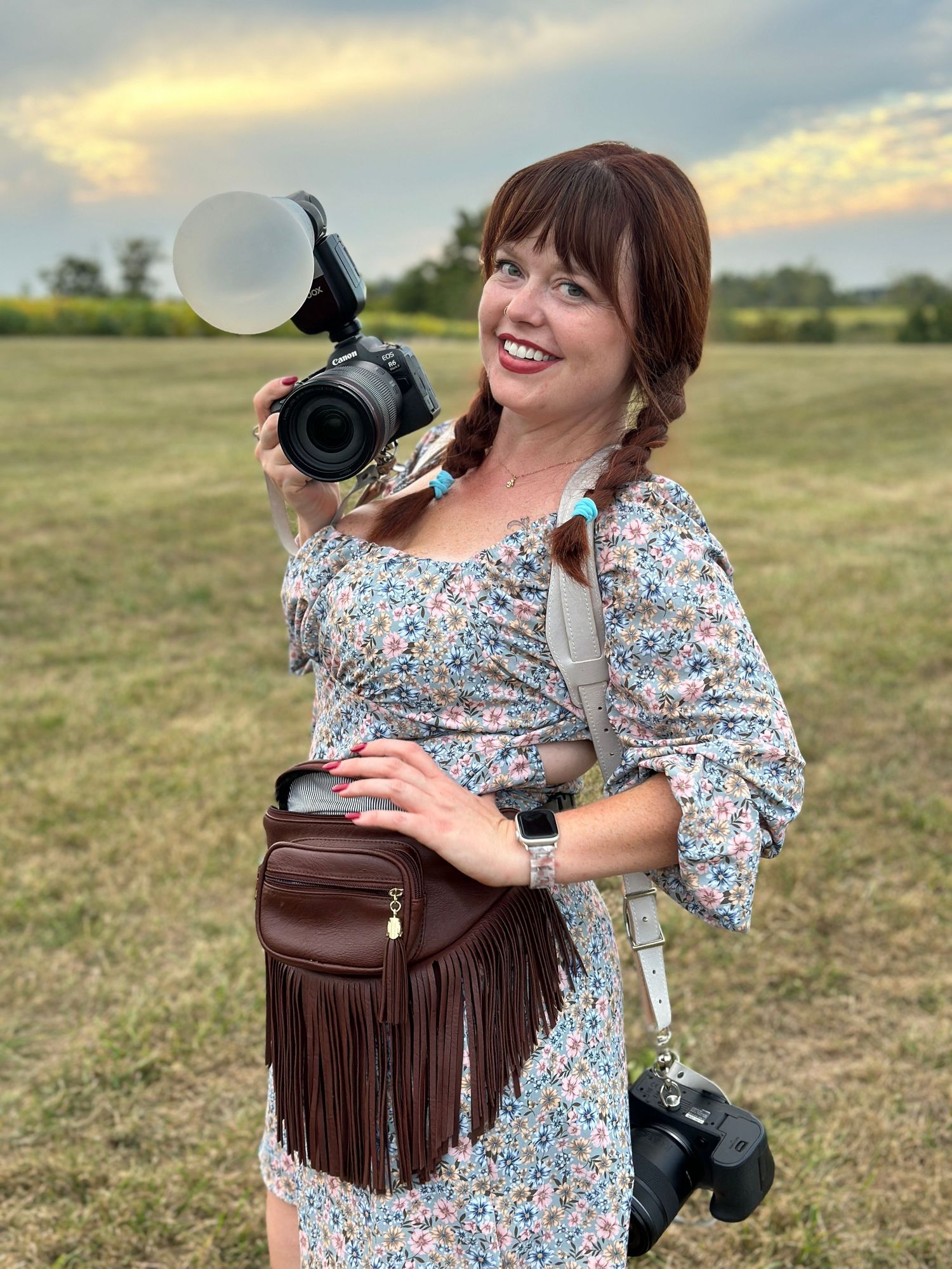 Smiling woman in floral dress holding a camera with flash diffuser in a green field at sunset.