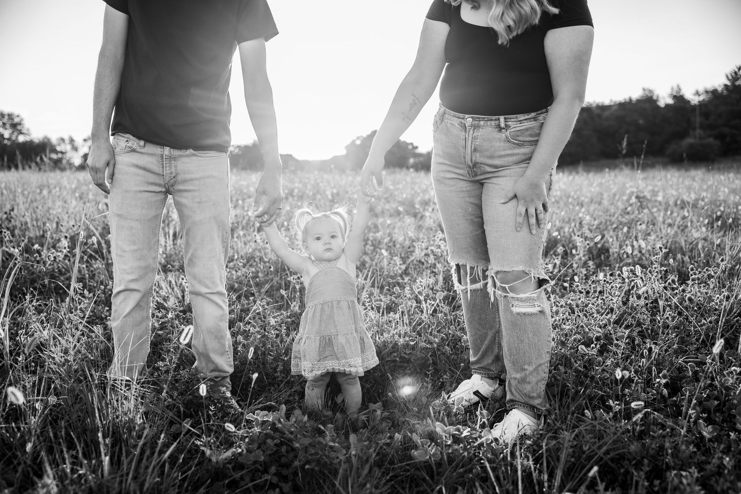 Black and white photo of parents holding hands with toddler in field at sunset.