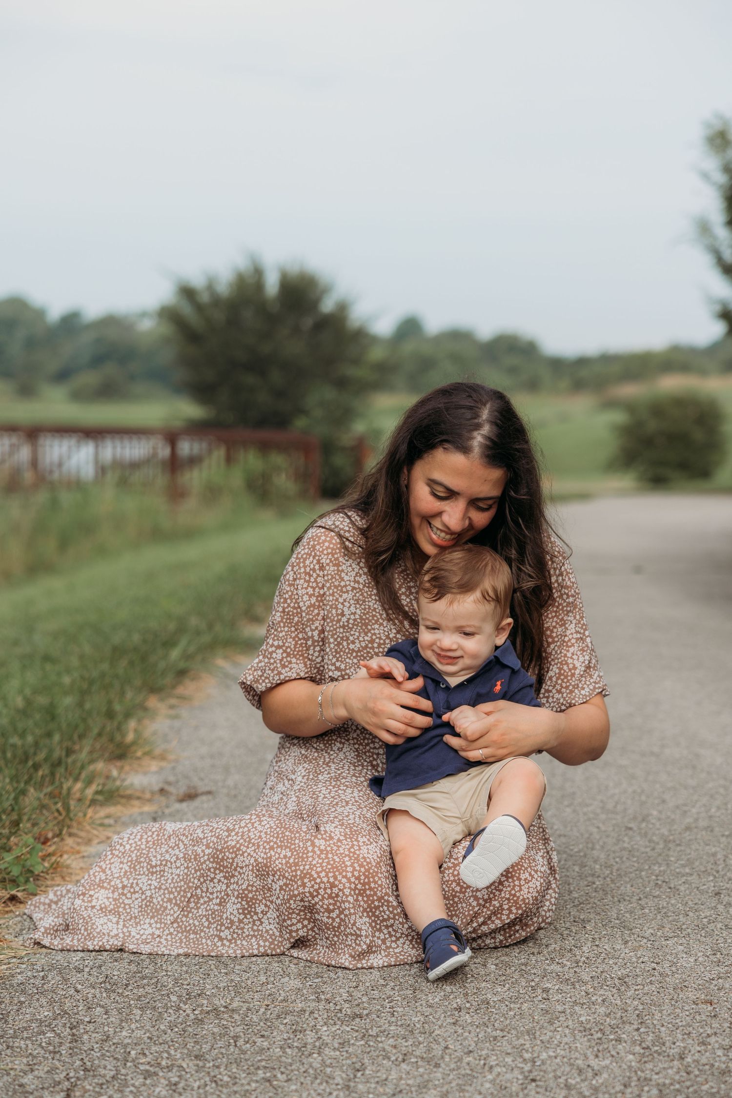 A smiling mother sits on a path cradling her toddler boy outdoors, surrounded by lush greenery.
