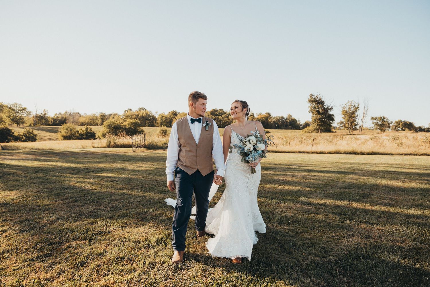 Bride and groom walking hand in hand across a sunlit green field in wedding attire, bride holding a white bouquet.