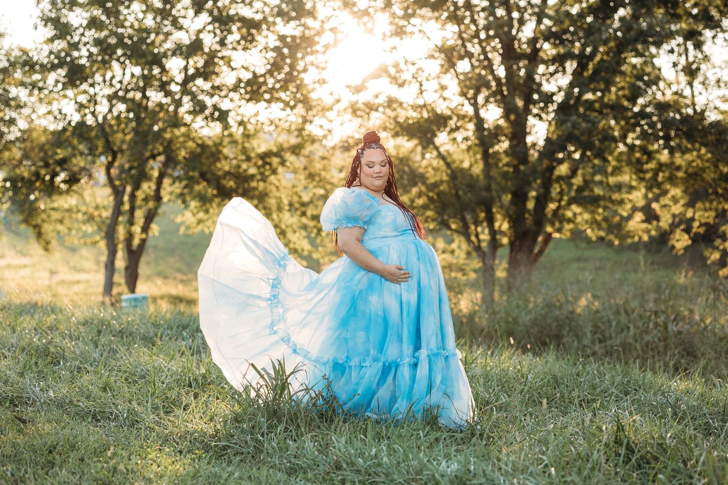 Pregnant woman in flowing light blue tulle gown poses outdoors in sunlit green field surrounded by trees.