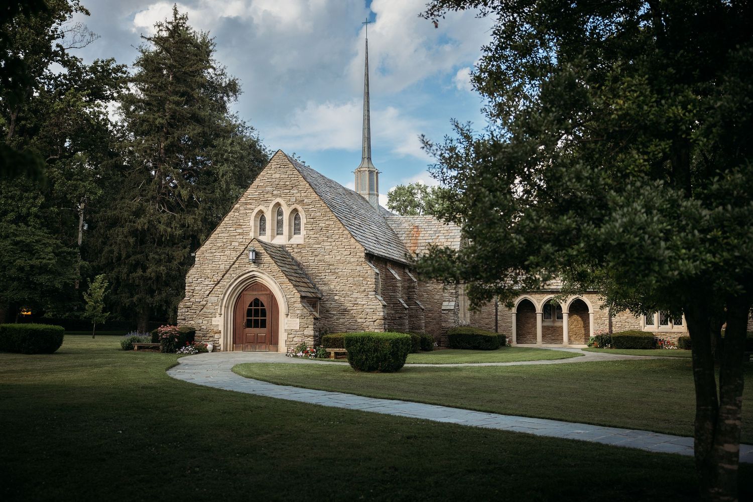 Historic stone church with Gothic architecture, arched wooden door, tall steeple, and lush green lawn surrounded by mature trees.