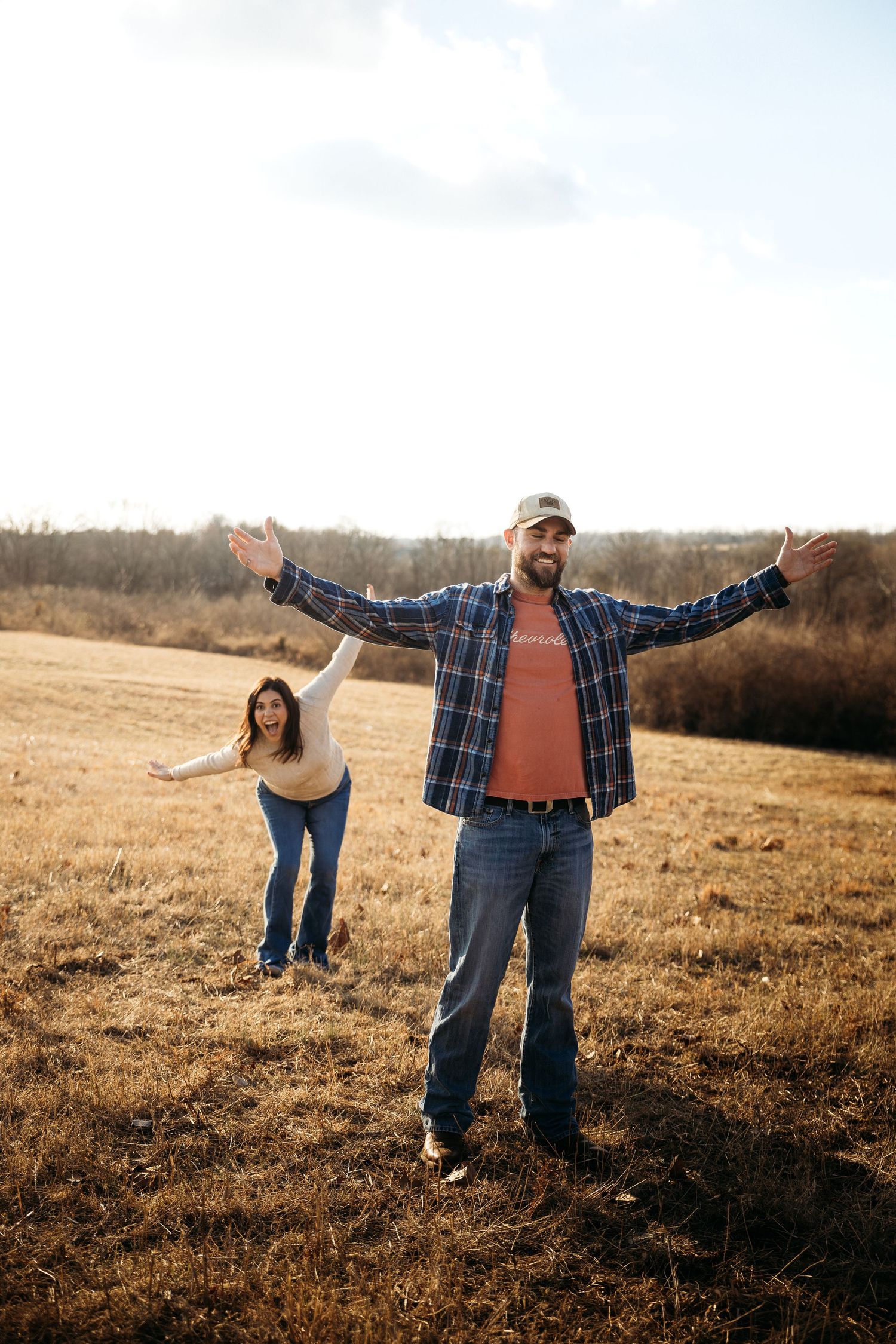 A joyful couple with arms outstretched standing in a golden field, wearing casual jeans and flannel on a sunny day.