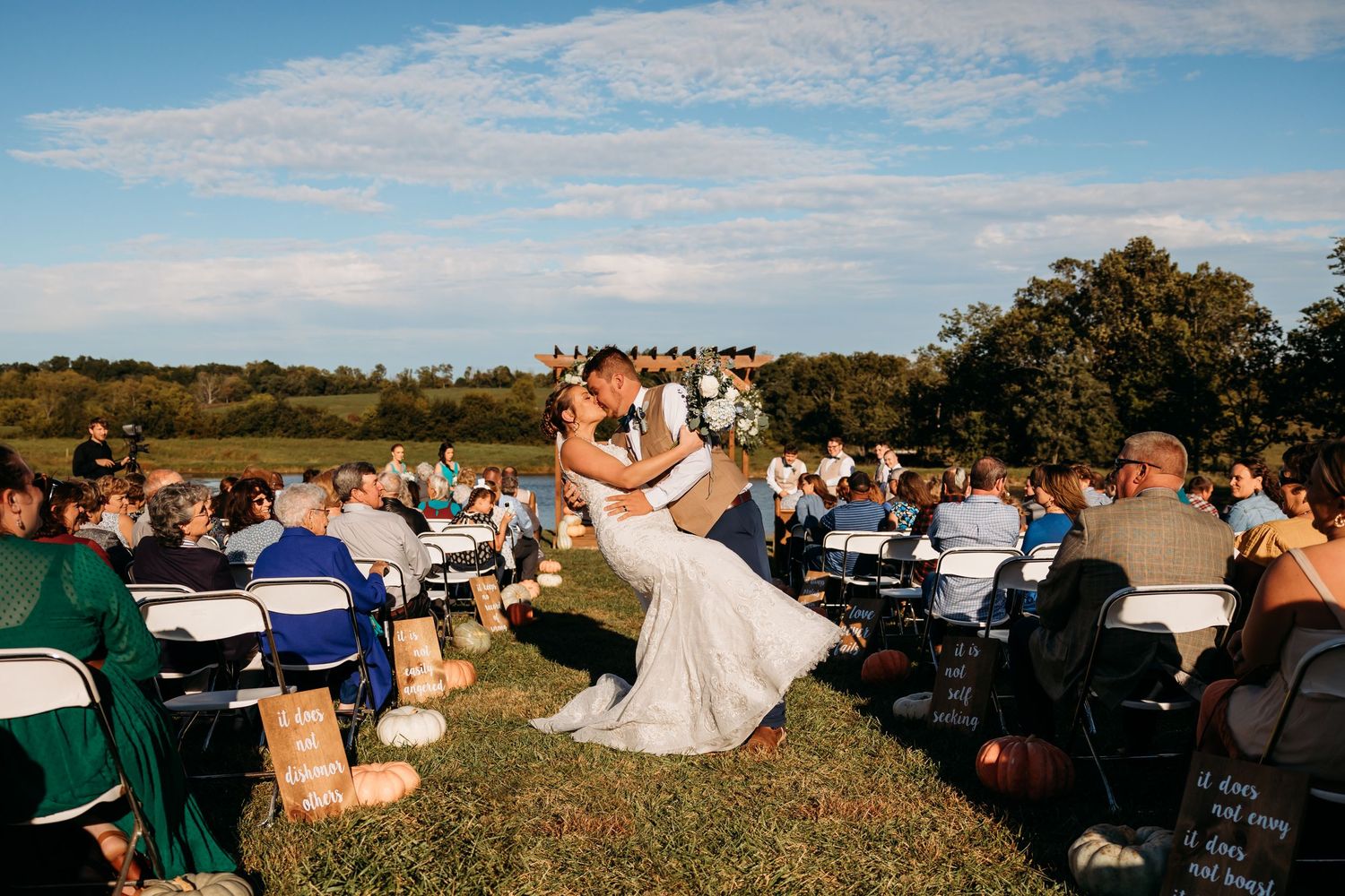 Bride and groom share a romantic kiss during an outdoor wedding ceremony on a sunny day with guests seated on white chairs.