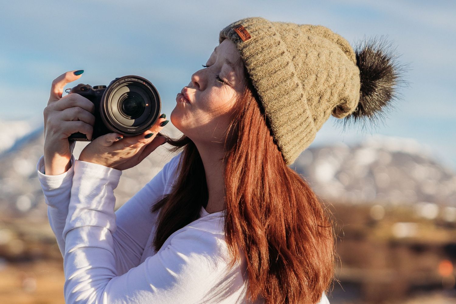 A young woman with red hair wearing a knit pom beanie photographs outdoors with a DSLR camera in a winter landscape.