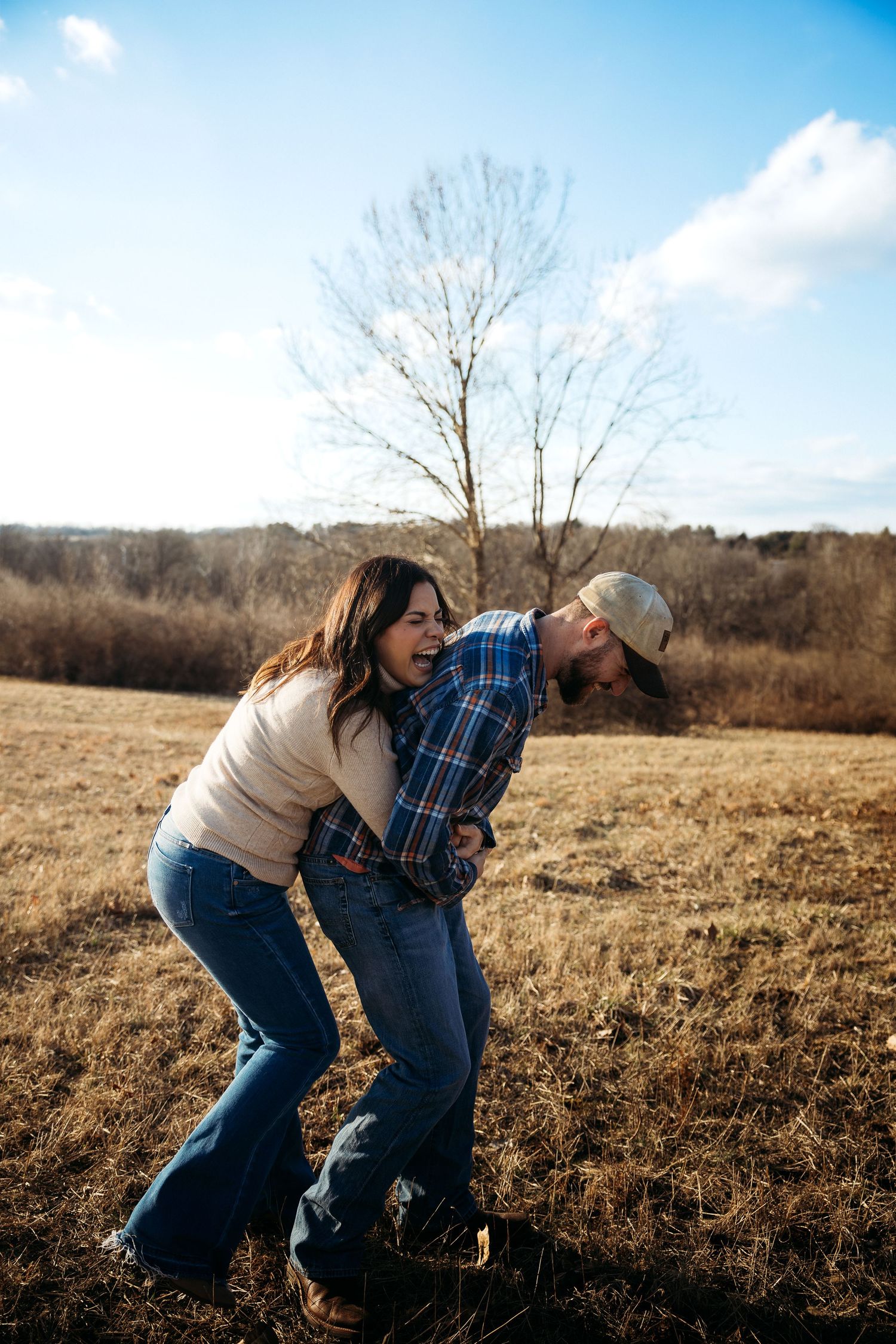 Couple laughing and embracing playfully in a golden field during autumn, with bare trees and blue sky behind them.