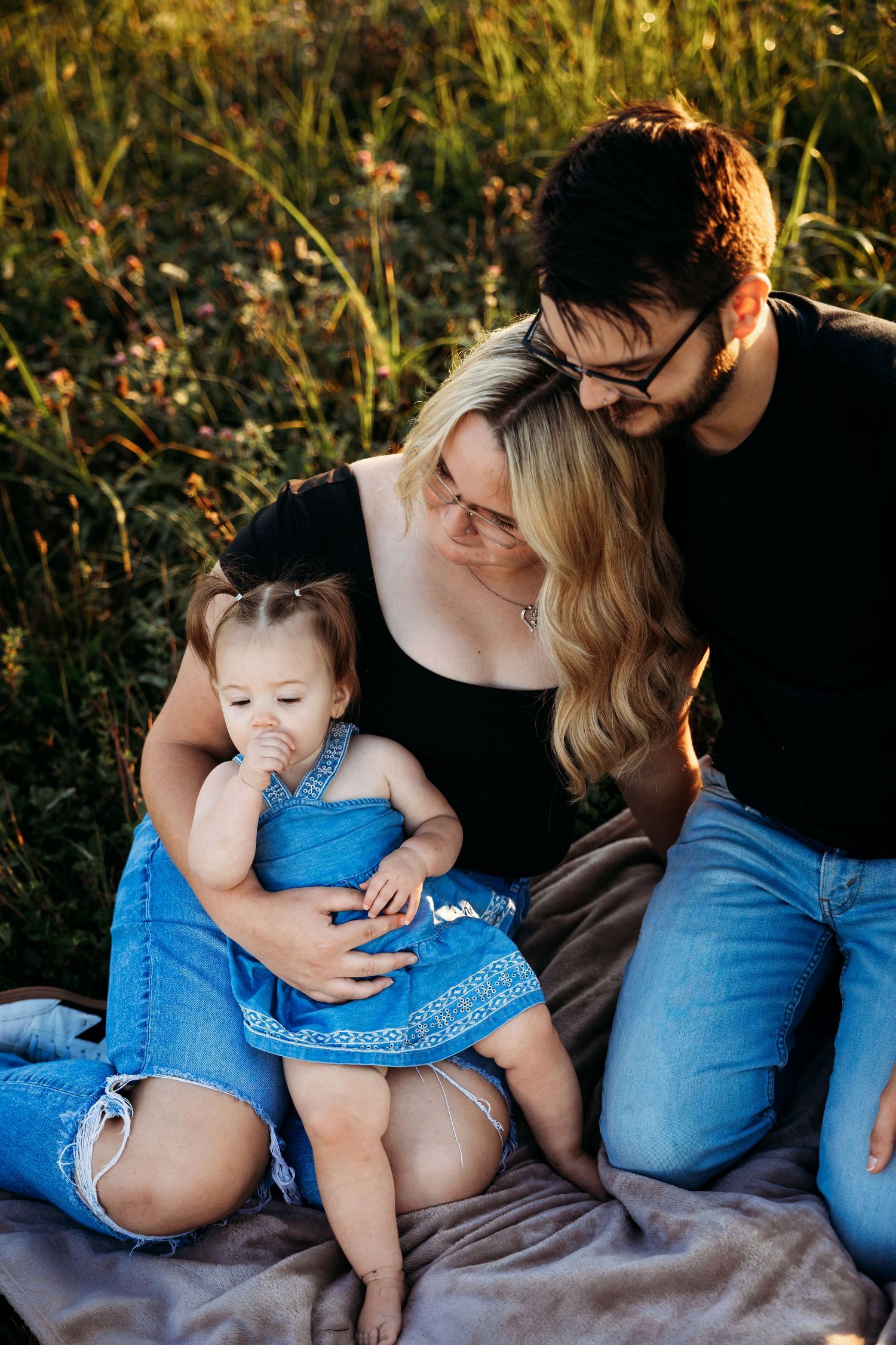 A young family sits on a blanket in a sunny field, parents smiling at their toddler girl in a blue denim dress.