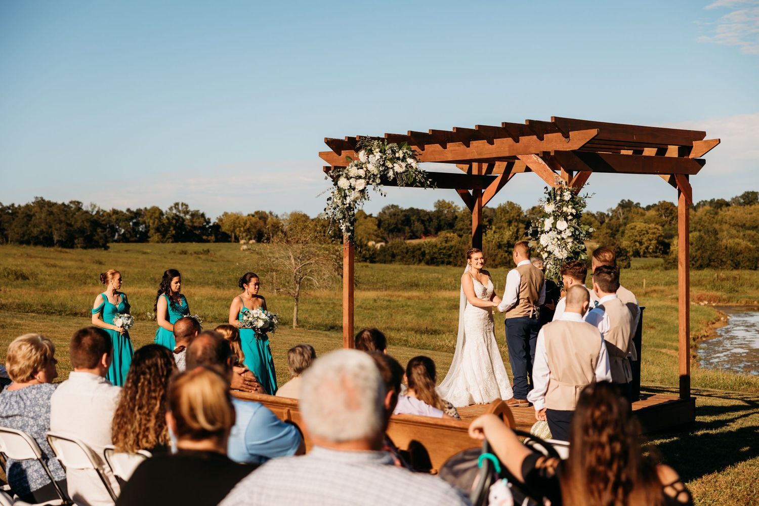 Outdoor wedding ceremony under a wooden pergola with floral decor, bridesmaids in teal, on a sunny green field.