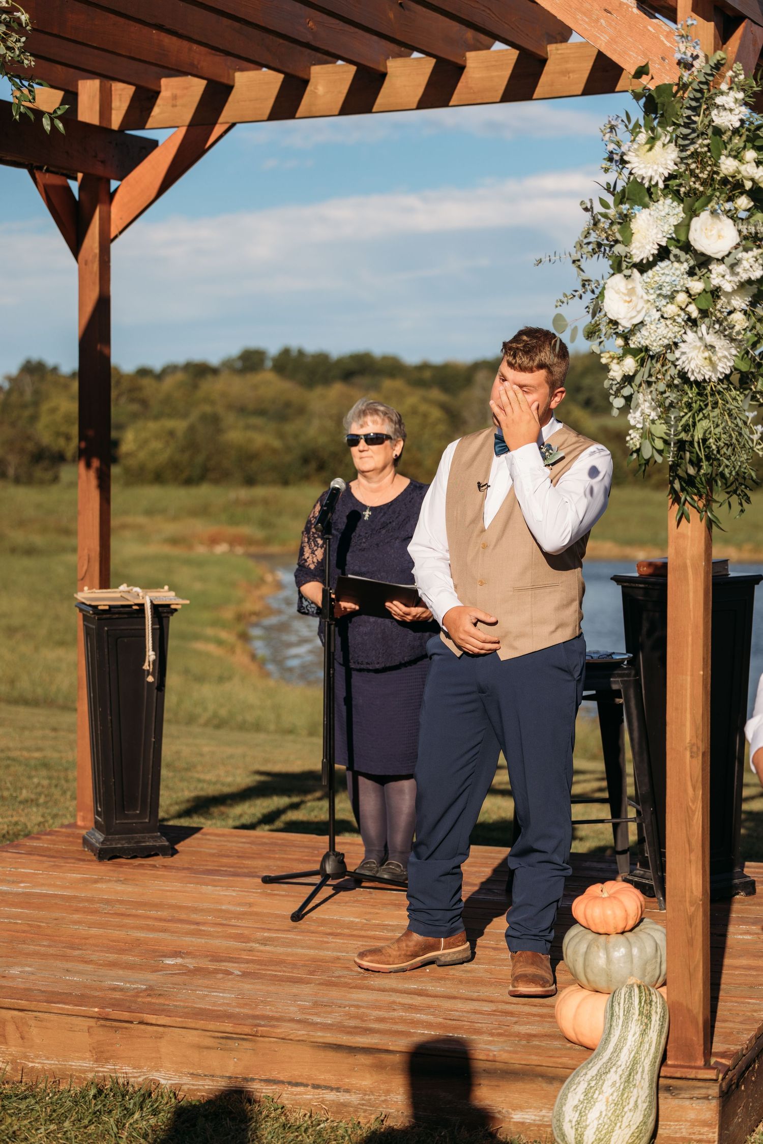 Emotional groom wiping tears at outdoor wedding ceremony under wooden pergola with white floral arrangements.