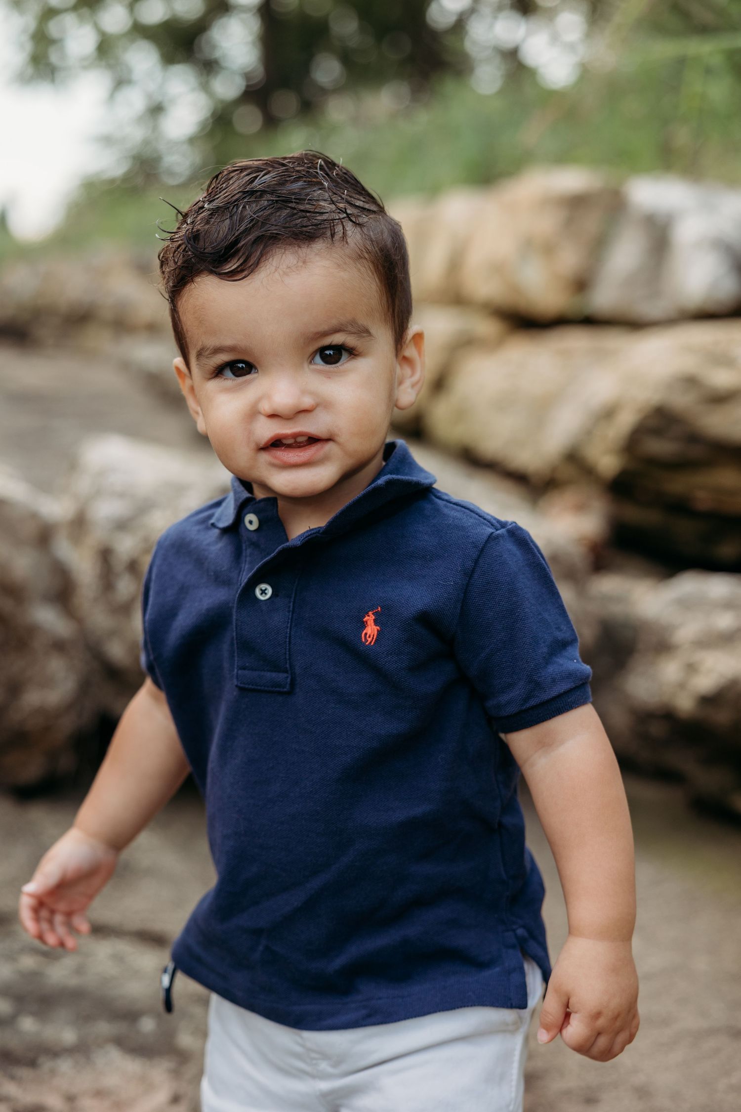 Adorable toddler boy in a navy Ralph Lauren polo shirt smiles outdoors near rocky terrain.