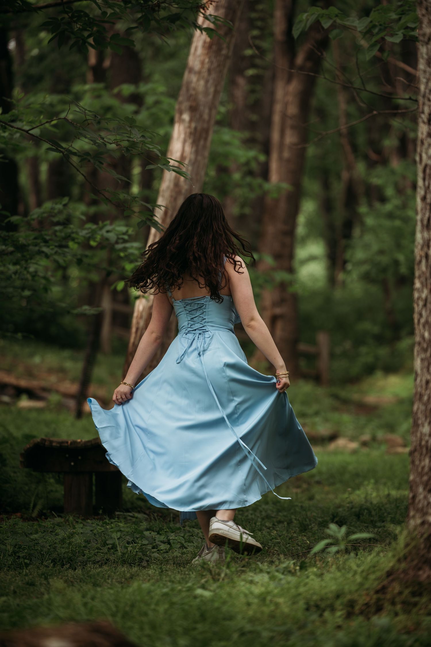 Woman in blue dress walking through lush green forest, viewed from behind with flowing skirt and dark hair.