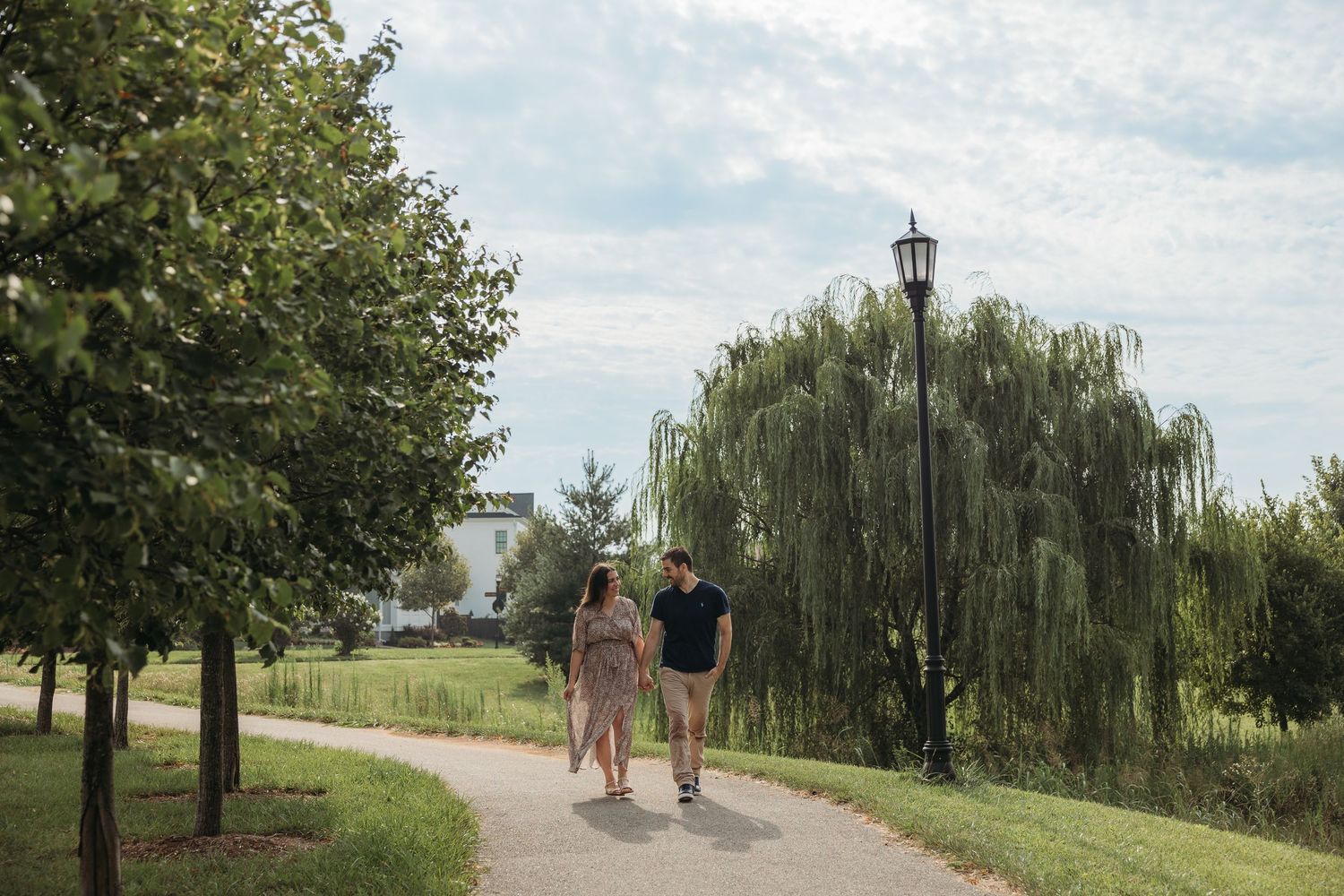 Couple holding hands walking on a curved park path lined with trees and a vintage lamp post on a cloudy day.