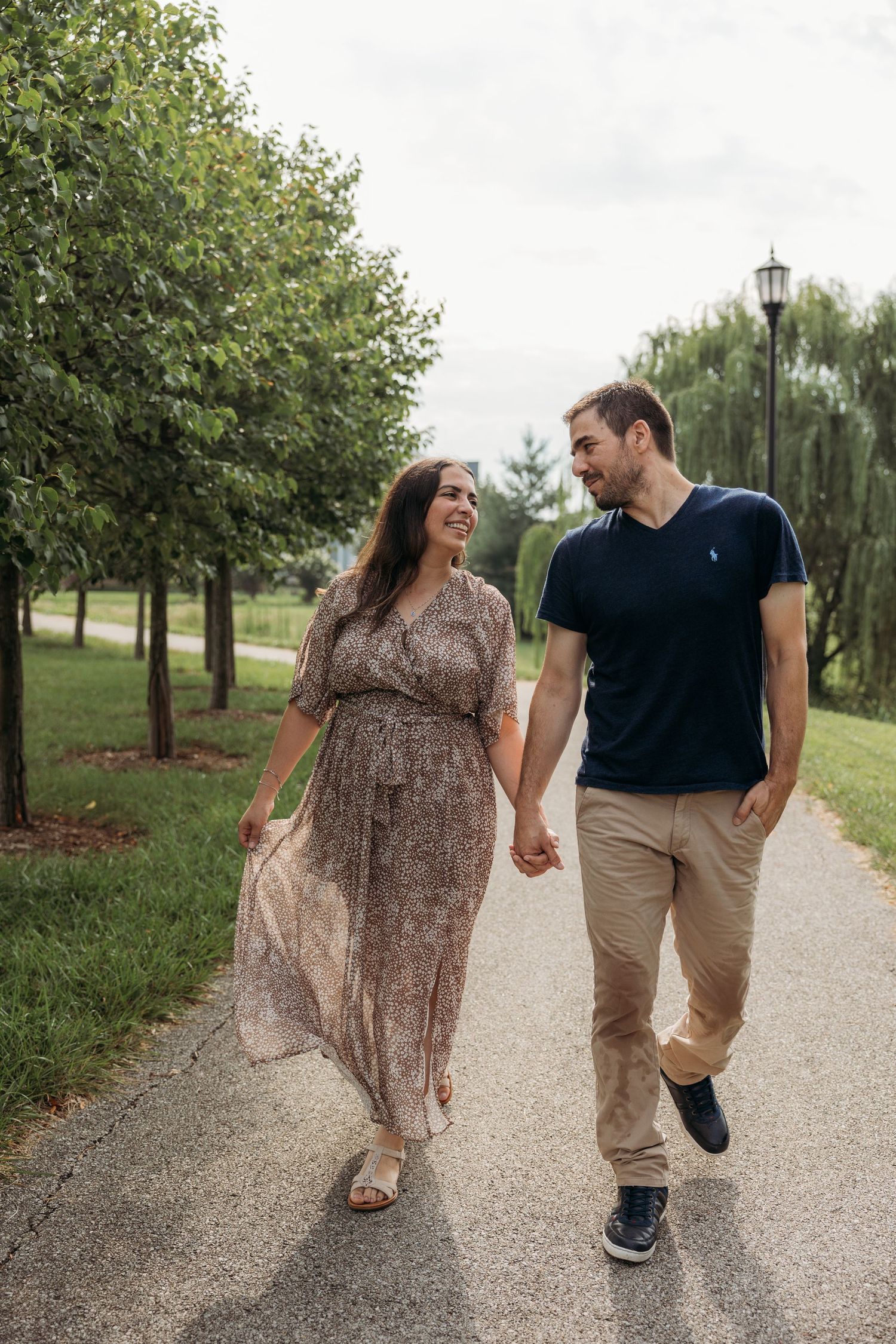 A couple holds hands and smiles at each other while walking along a park path surrounded by green trees.