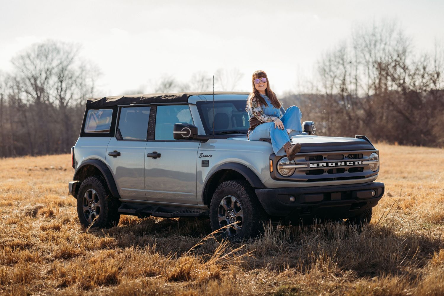 Woman in blue jeans sitting on hood of silver Ford Bronco SUV in a golden field during autumn.
