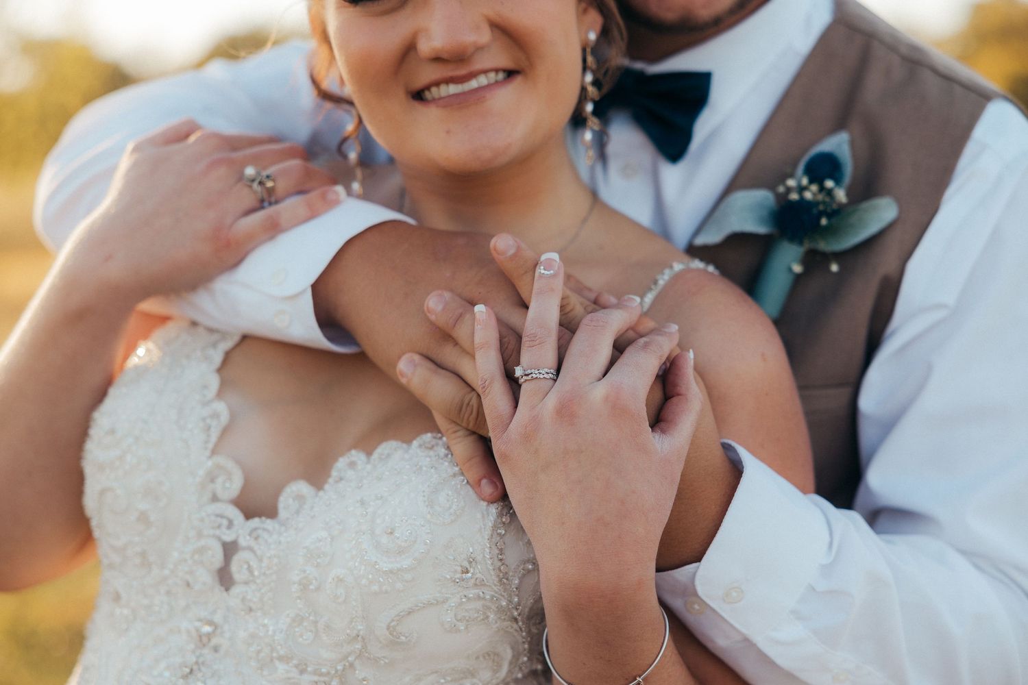 Close-up of bride and groom holding hands showing wedding rings in lace wedding dress and tan vest.