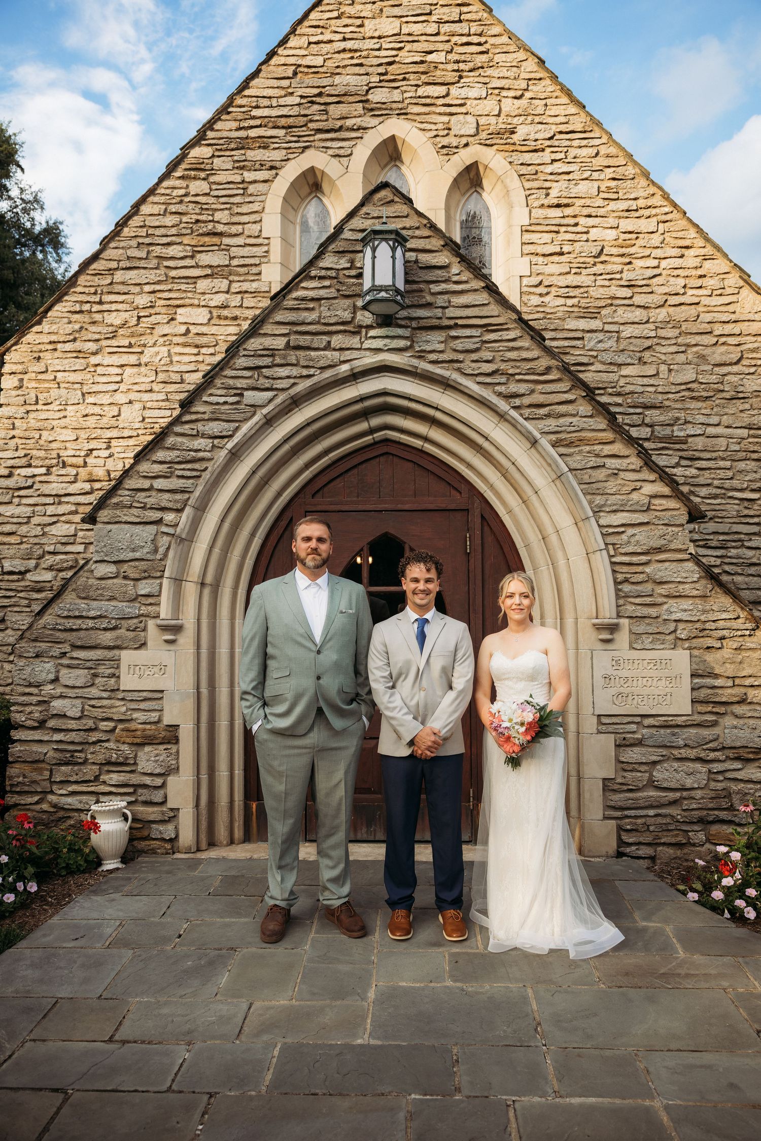 Bride and groom pose with officiant before stone church Gothic arch entrance on wedding day.
