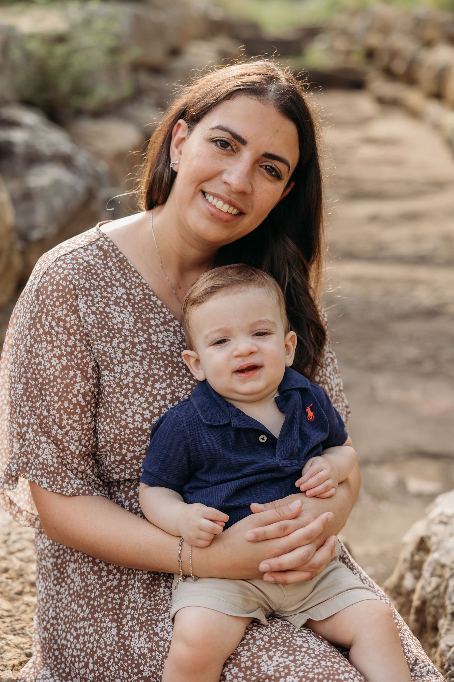 A smiling mother holds her baby boy on her lap while sitting outdoors on rocky steps.