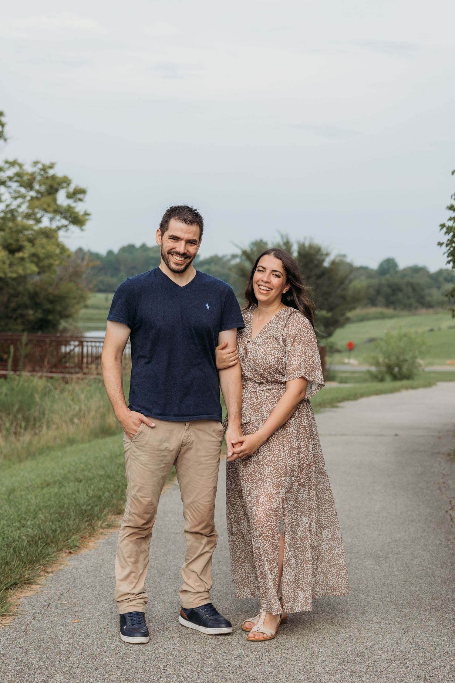 Smiling couple holding hands on a scenic path, man in navy polo and woman in floral maxi dress.