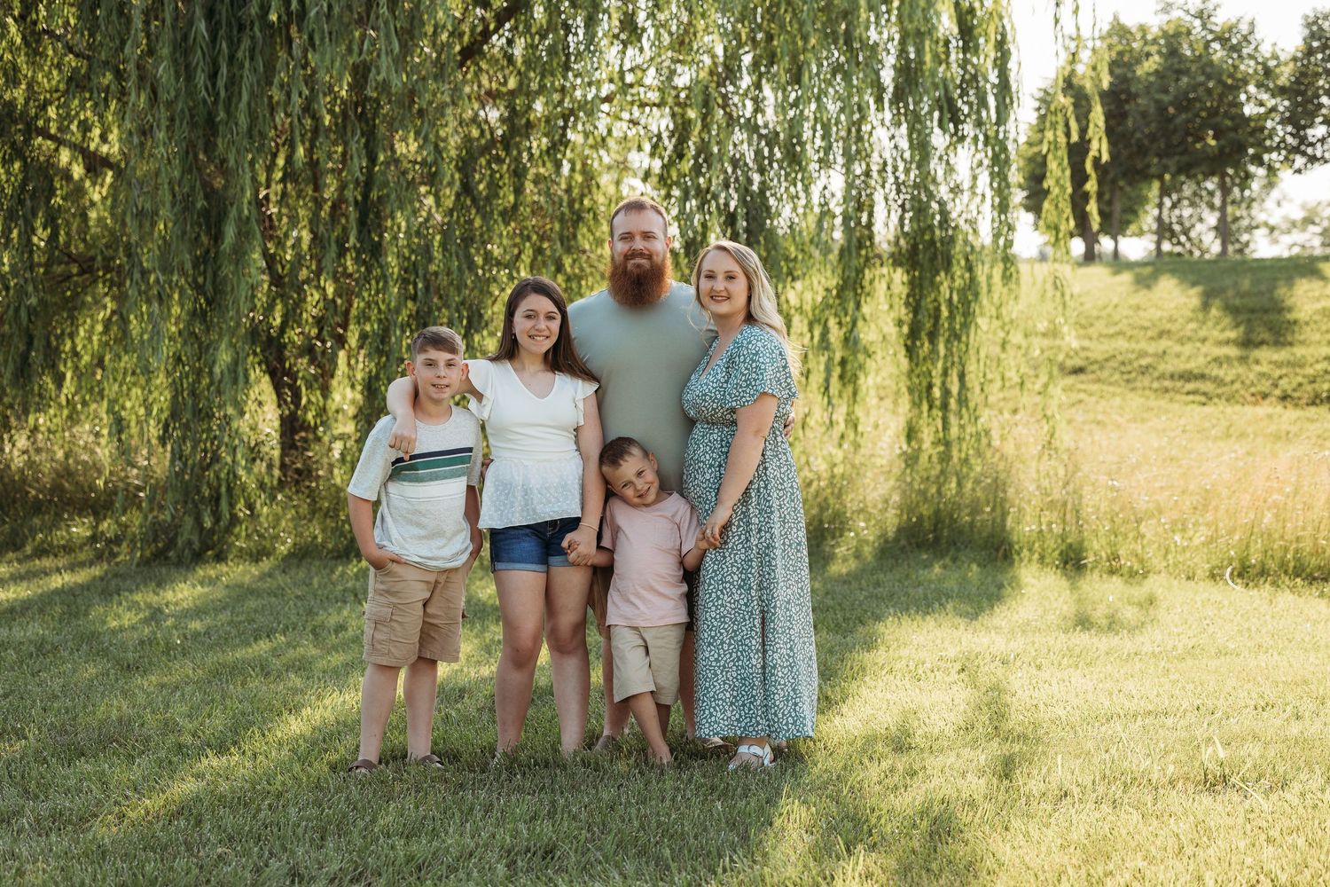 Family of five smiling outdoors under a weeping willow tree on a sunny day in a green field.