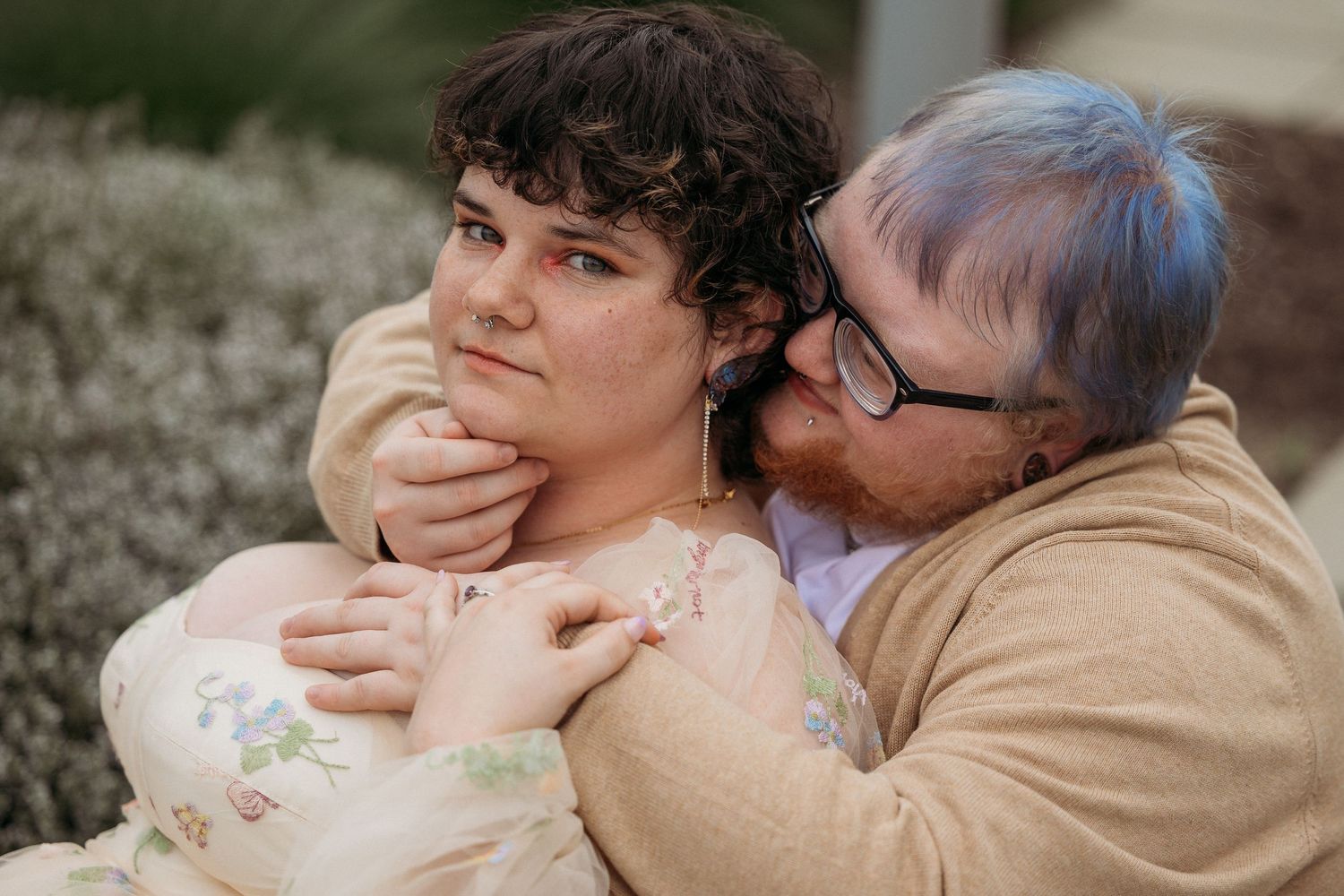 Couple sharing intimate moment outdoors, man in tan cardigan embracing woman in floral dress, kissing her cheek.