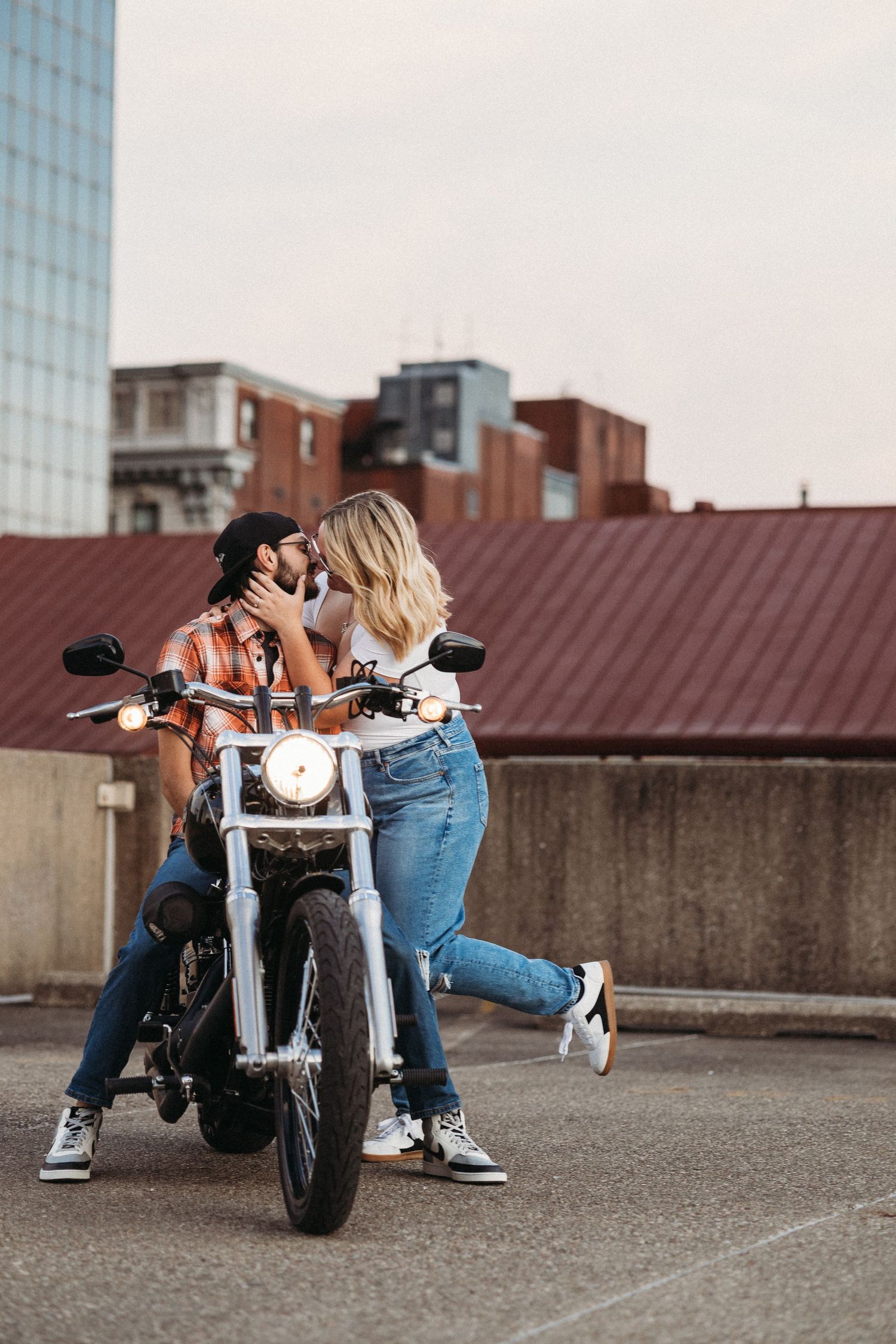 Romantic couple kissing beside a motorcycle on a rooftop parking lot with urban buildings in the background.