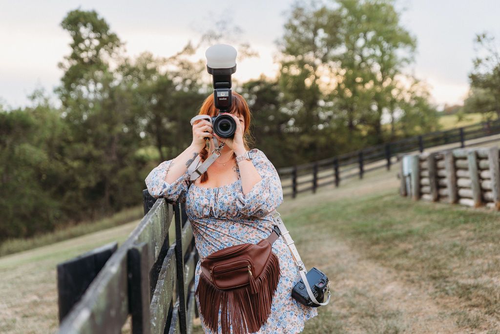 Woman in floral dress photographing with DSLR camera outdoors near wooden fence with green trees in background.